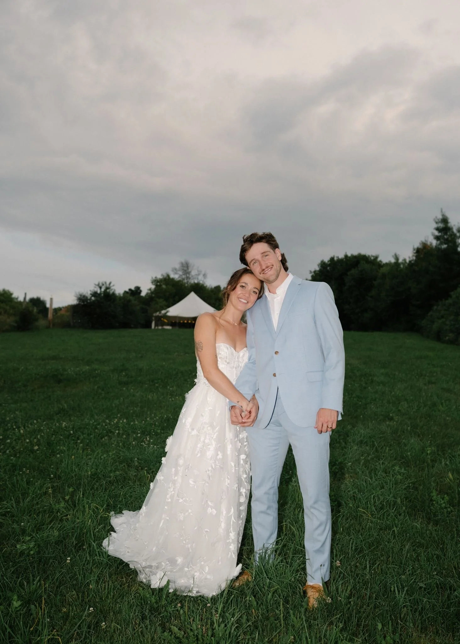 Un couple en habits de mariage souriant dans un champ vert avec un ciel nuageux, un chapiteau blanc en arrière-plan.