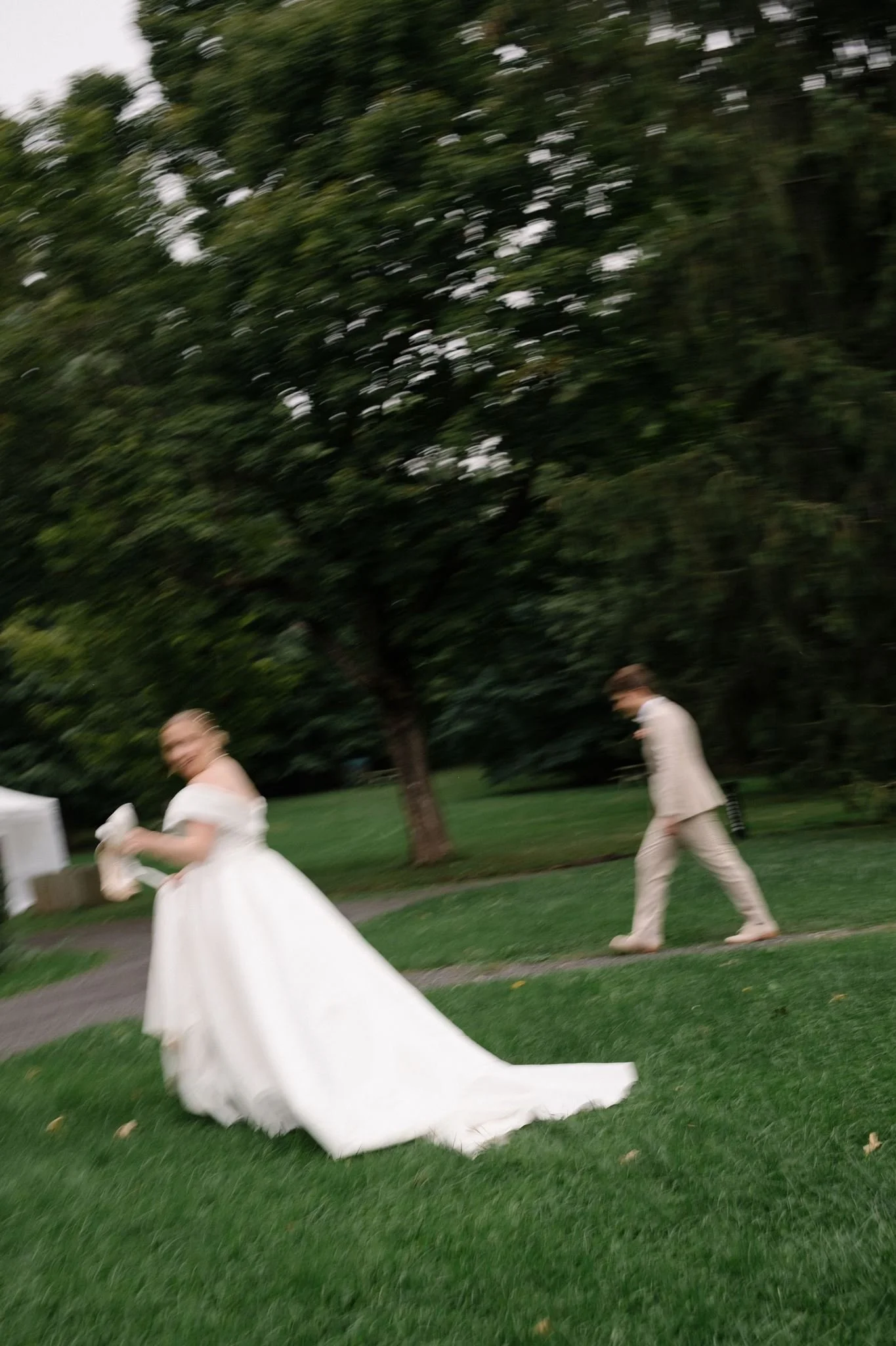 Une femme en robe blanche lors d'un mariage qui marche sur une pelouse, avec un homme en costume beige qui marche derrière elle, dans un parc avec des arbres.