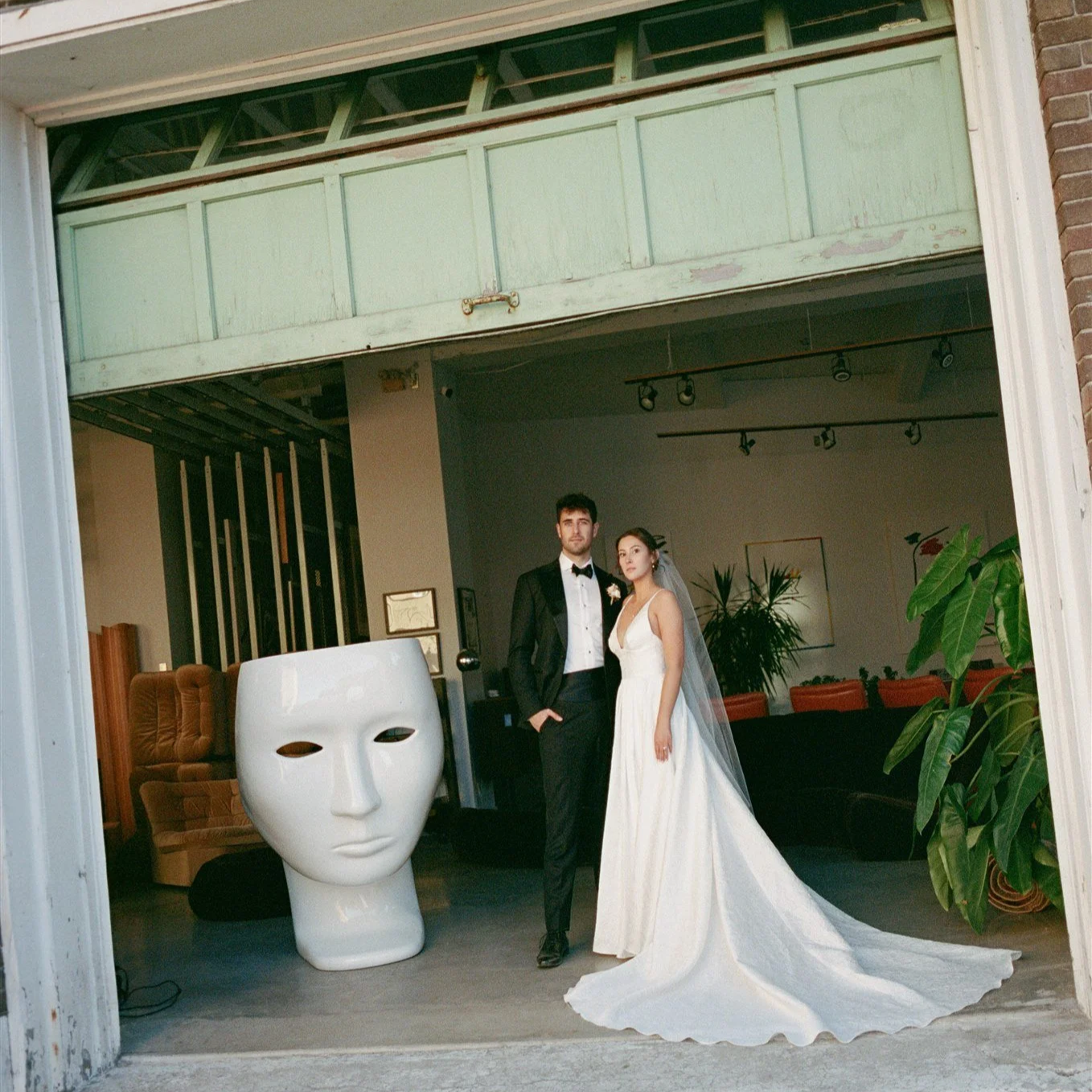Un couple en costume de mariage posant dans un espace intérieur avec des plantes vertes et des meubles modernes, vu à travers une ouverture de garage ou d'atelier. Une sculpture en forme de visage blanc est visible à gauche.