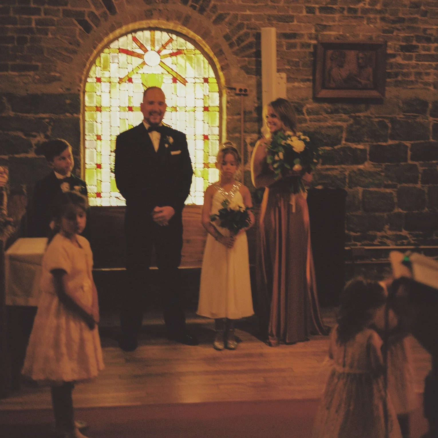 Une cérémonie de mariage dans une église en pierre avec un vitrail coloré en arrière-plan, où le marié, la mariée et plusieurs enfants sont présents.