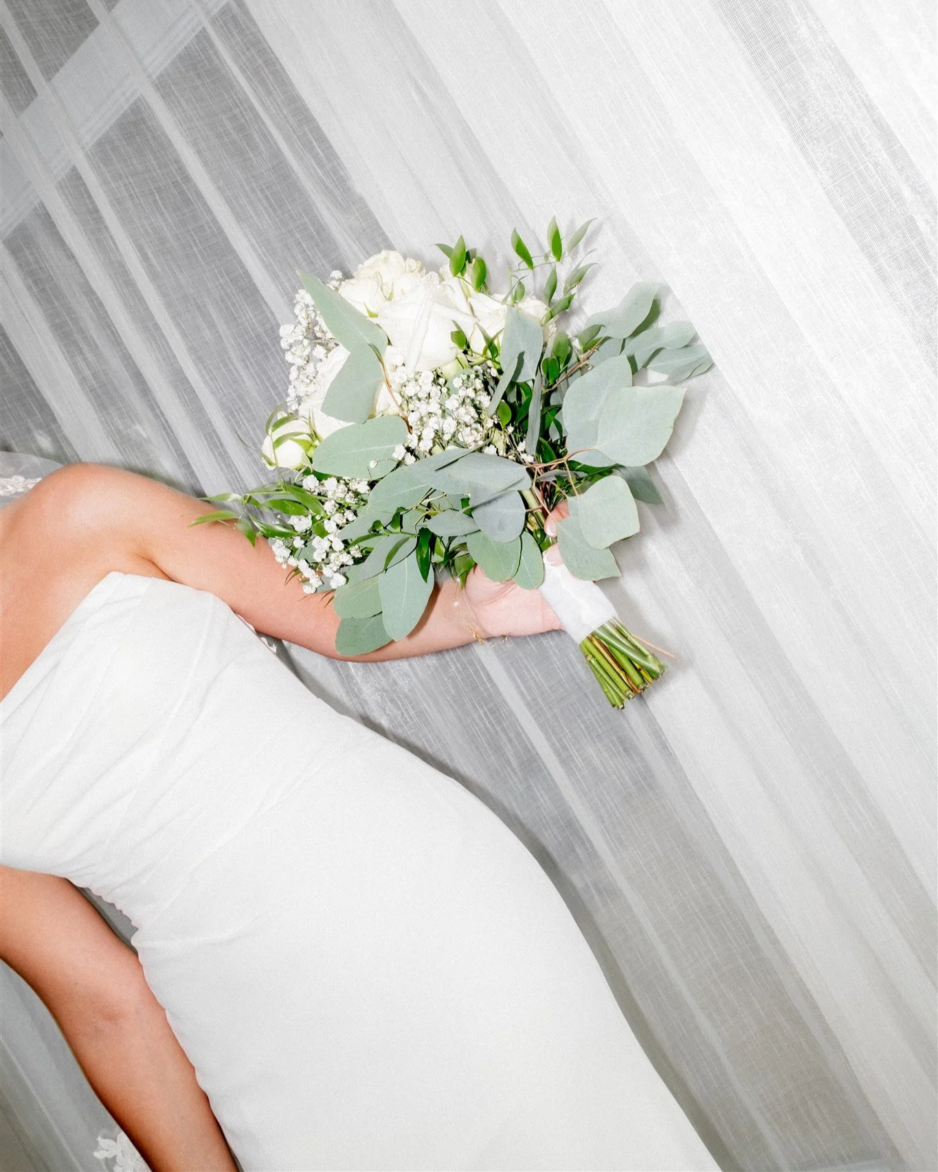 Un bouquet de fleurs blanches et vertes est tenu par une personne portant une robe blanche, posé sur un lit avec une porte en arrière-plan.
