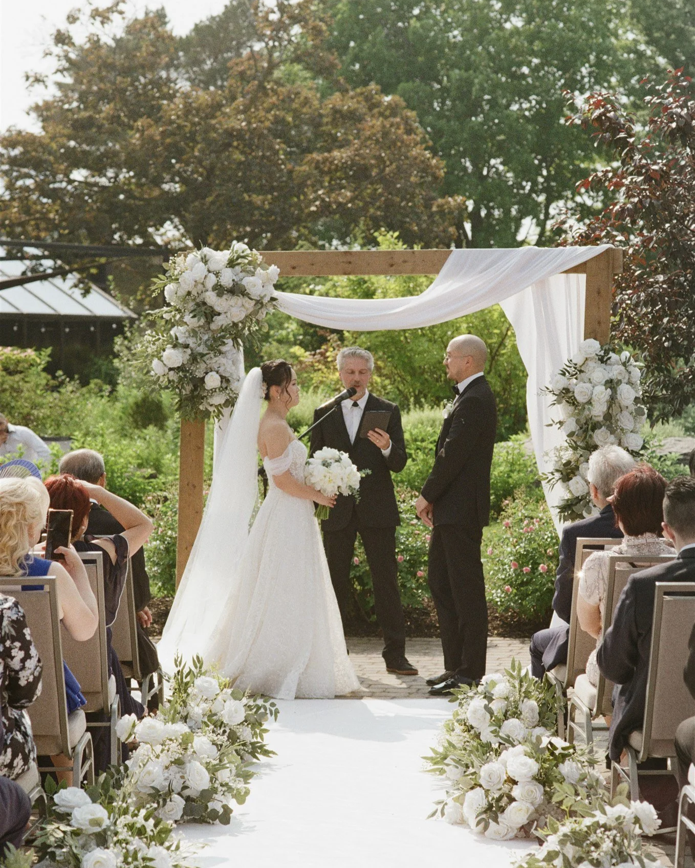 Cérémonie de mariage en plein air avec un couple échangeant leurs vœux sous un arc décoré de fleurs blanches et de tissu blanc, entouré d'invités assis et d'une végétation luxuriante.