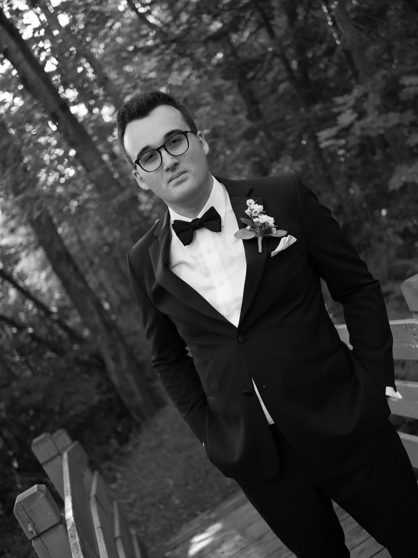 Un homme en costume noir avec une chemise blanche, une cravate noire et une boutonnière florale pose dans un parc en forêt.