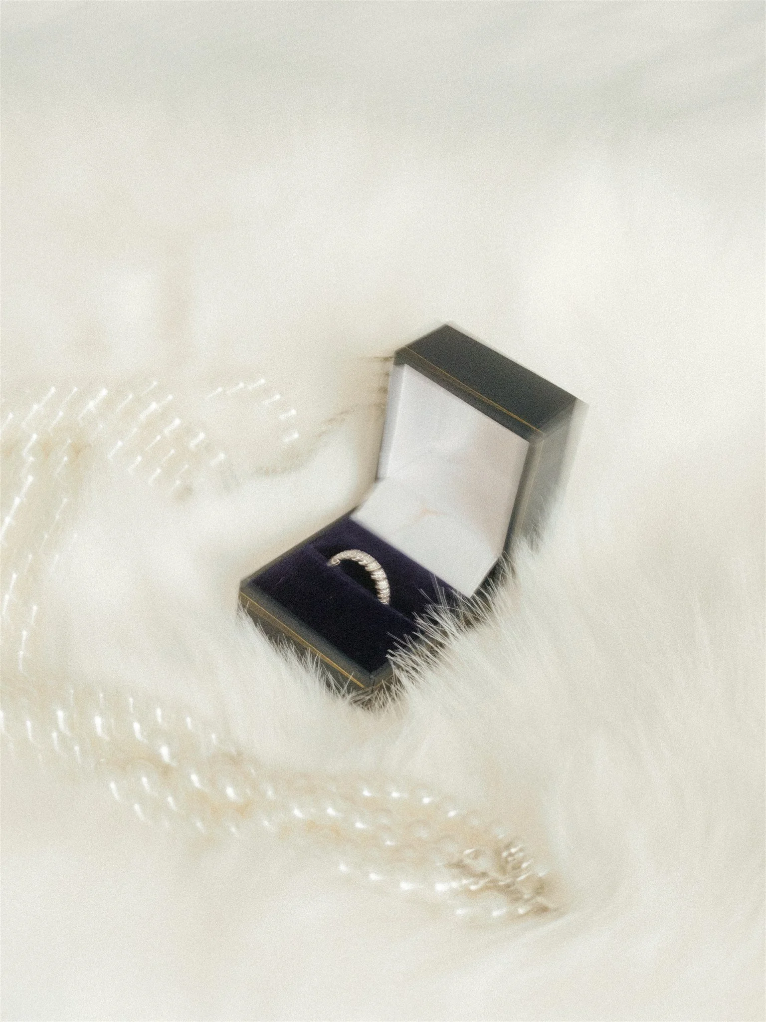 Une boîte noire contenant une bague en argent, posée sur un tissu blanc ou crème avec un motif de détails faits de points ou de petits trous.