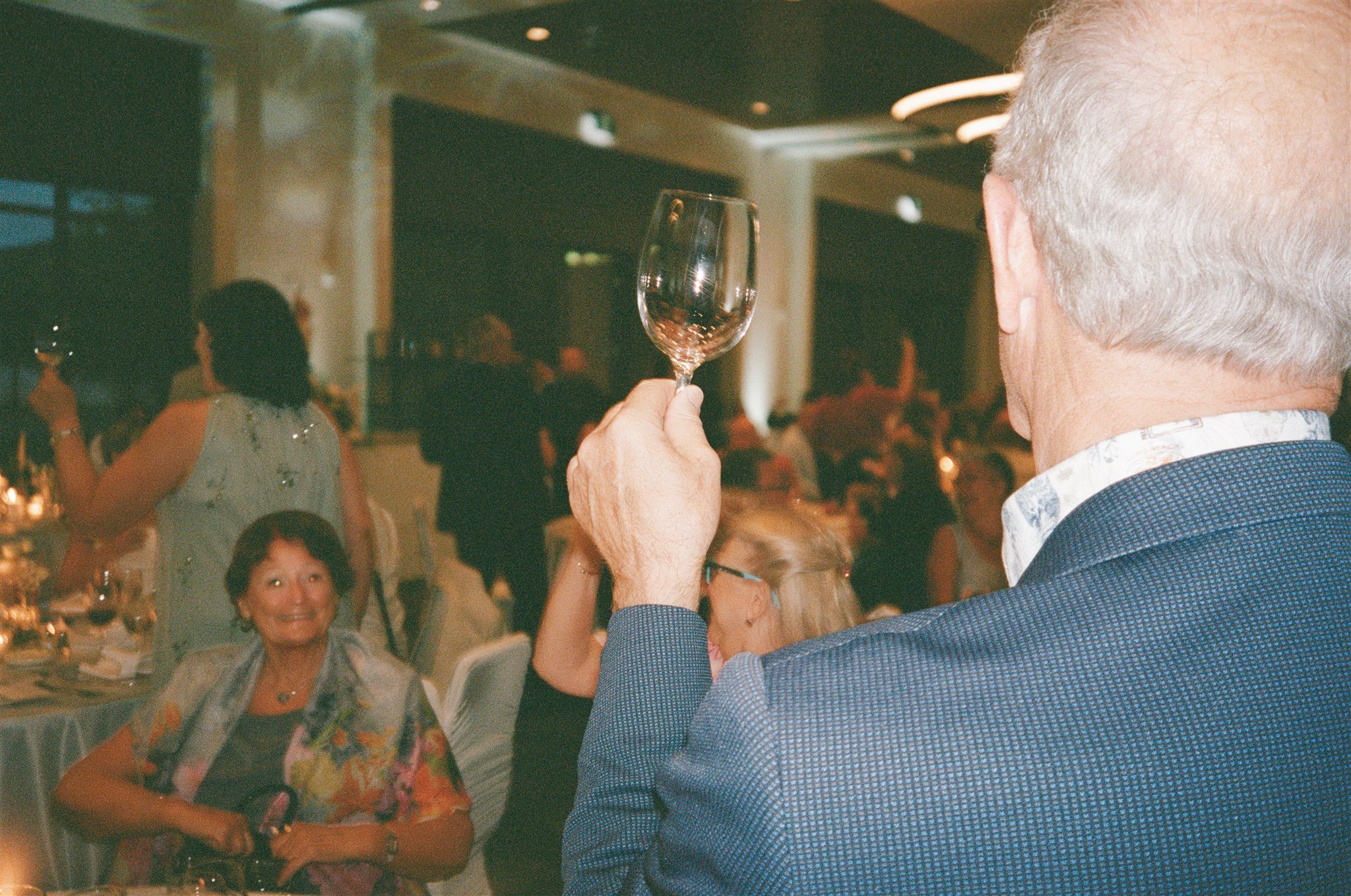 Un homme âgé en costume tient un verre à vin sans liquide dans un salon de fête, avec d'autres personnes souriantes en arrière-plan.