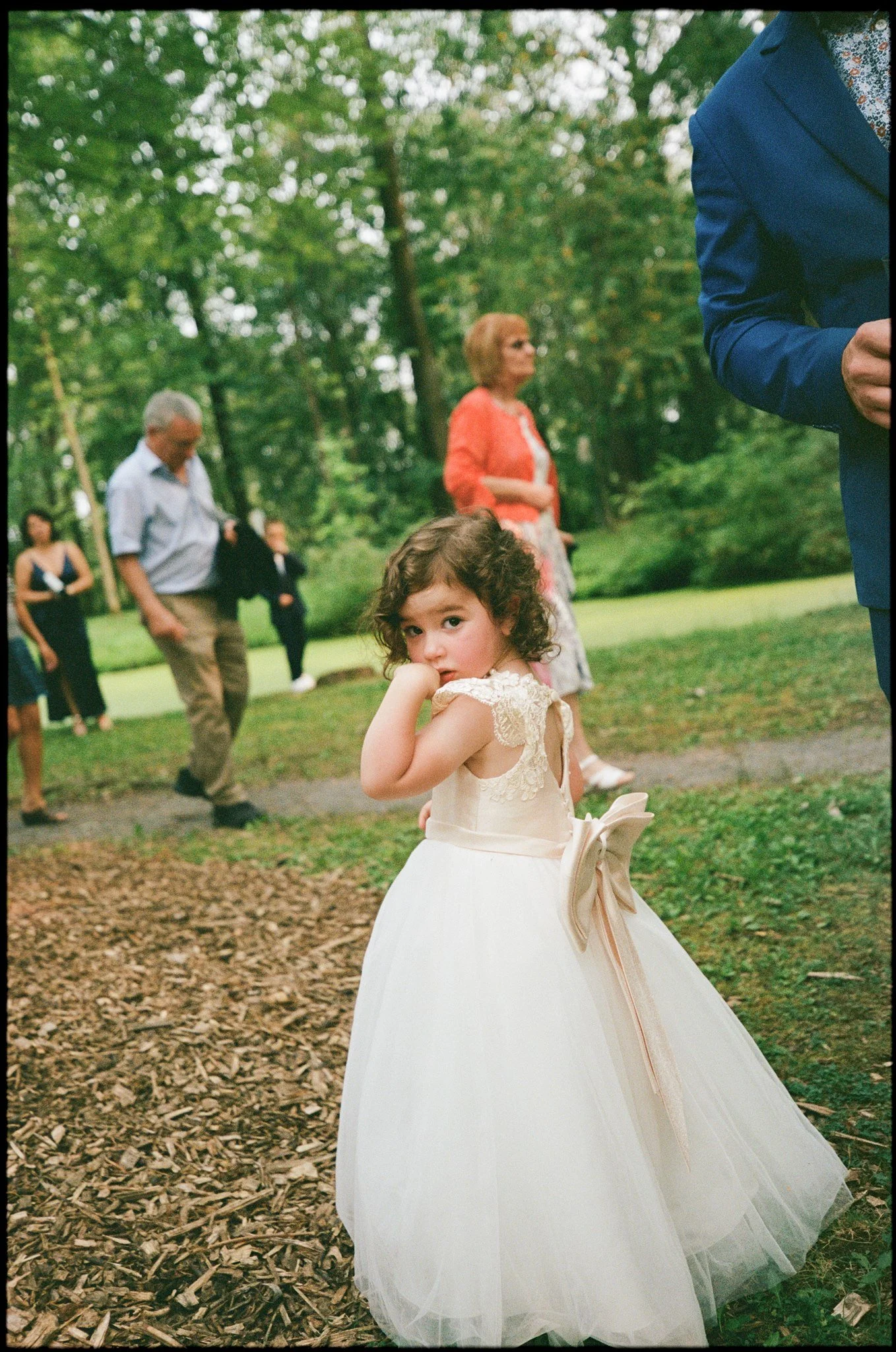 Une jeune fille en robe de mariée blanche avec un large ruban beige, posant dans un parc verdoyant lors d'un événement en plein air.