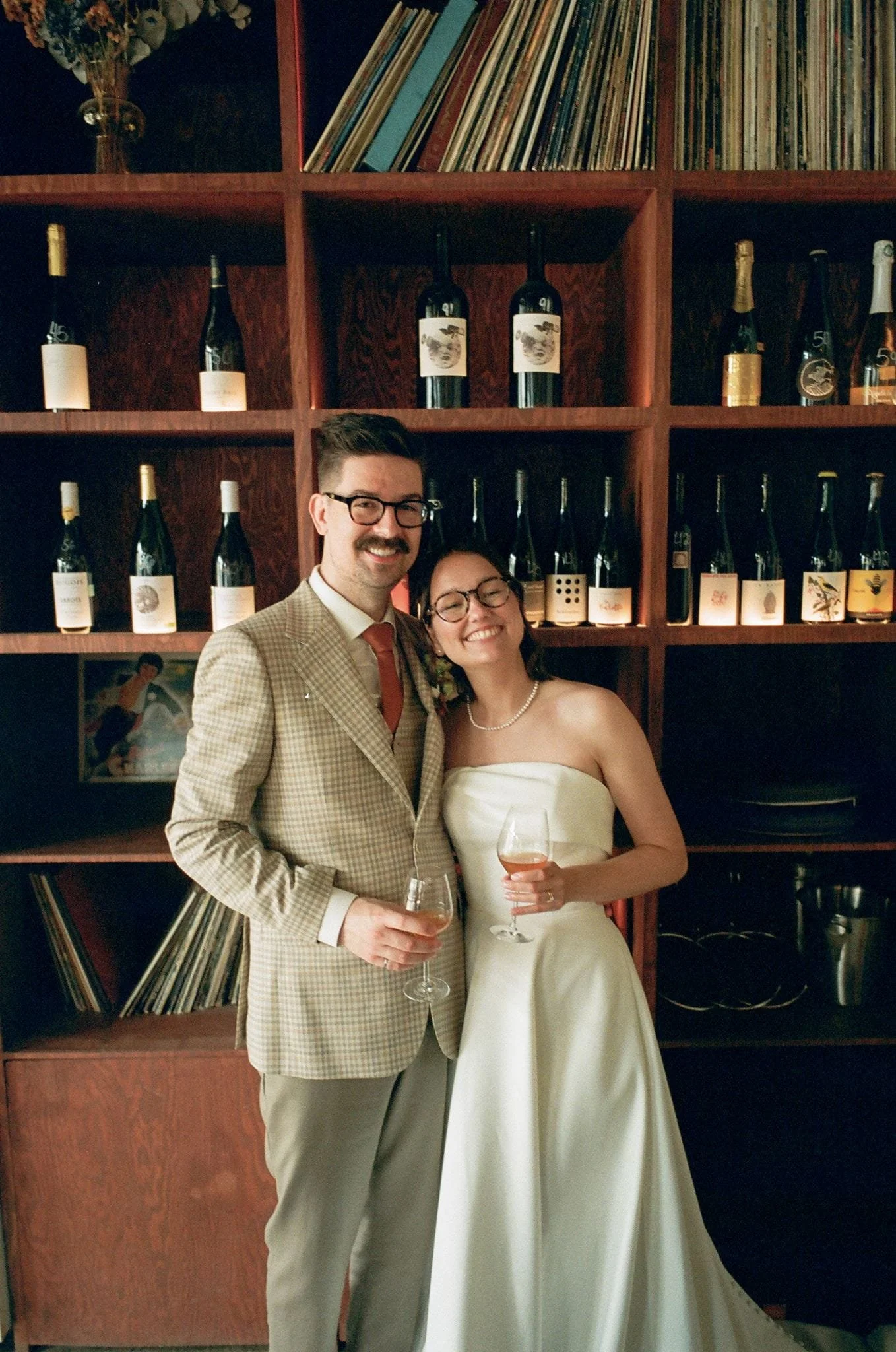 Un homme et une femme habillés de façon élégante, tenant chacun un verre de vin rose, posent devant une étagère remplie de vin en bouteille et de disques vinyles.