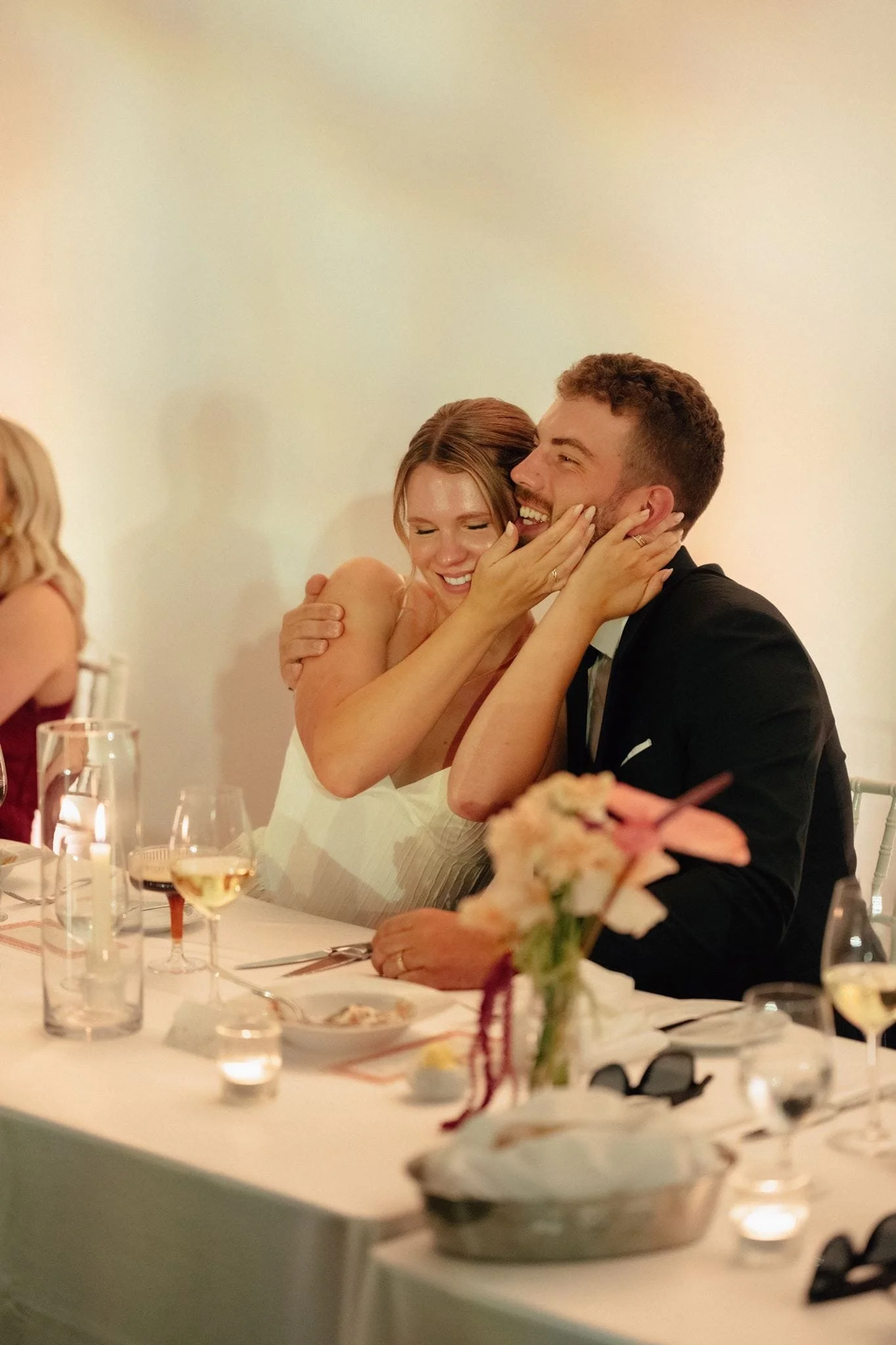 Un couple souriant lors d'une célébration ou d'un mariage, assis à une table avec des verres et des fleurs décoratives.