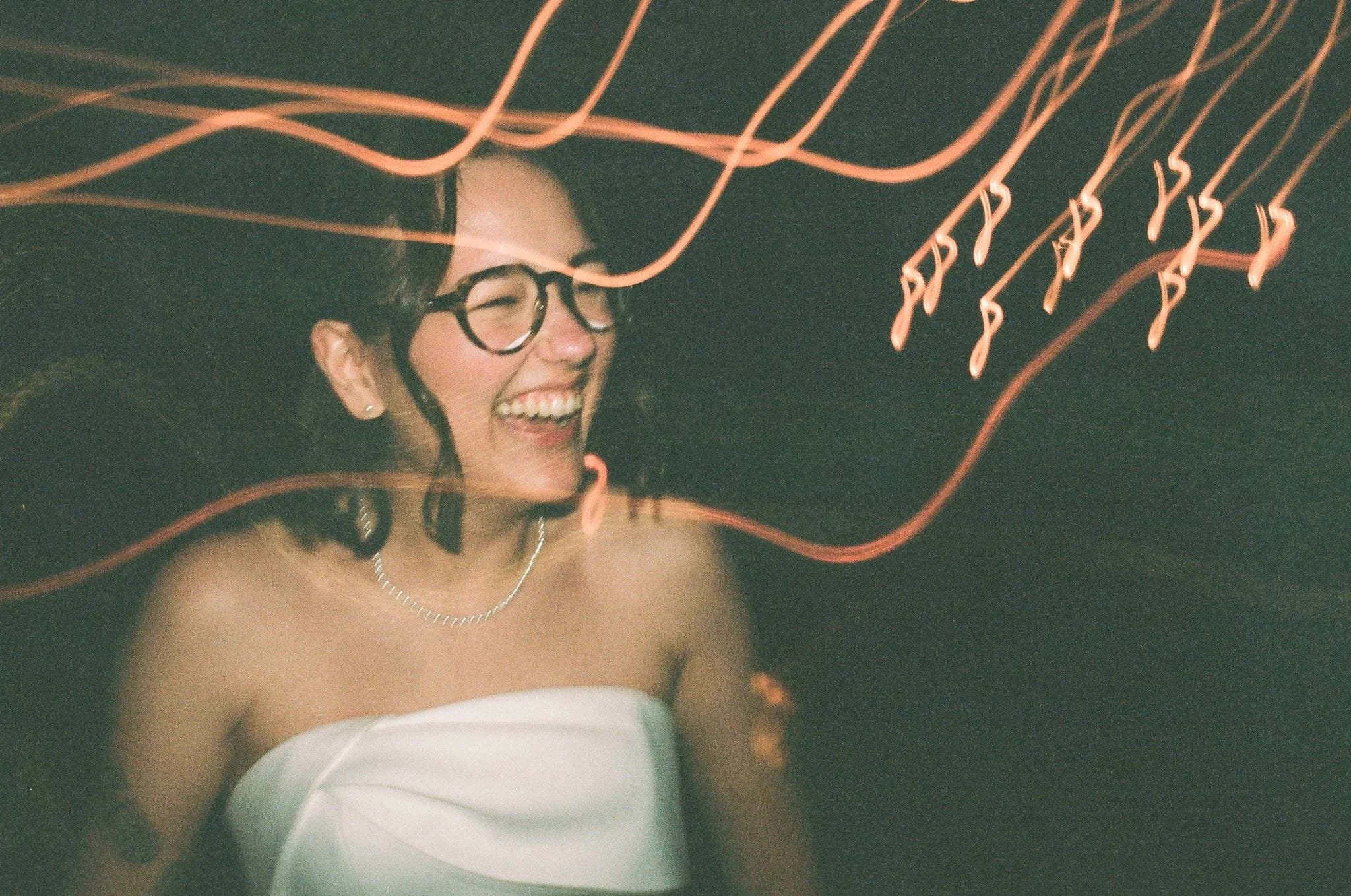 Femme souriante avec lunettes, portant un collier, dans un environnement sombre avec des lumières rouge orangé en arrière-plan.