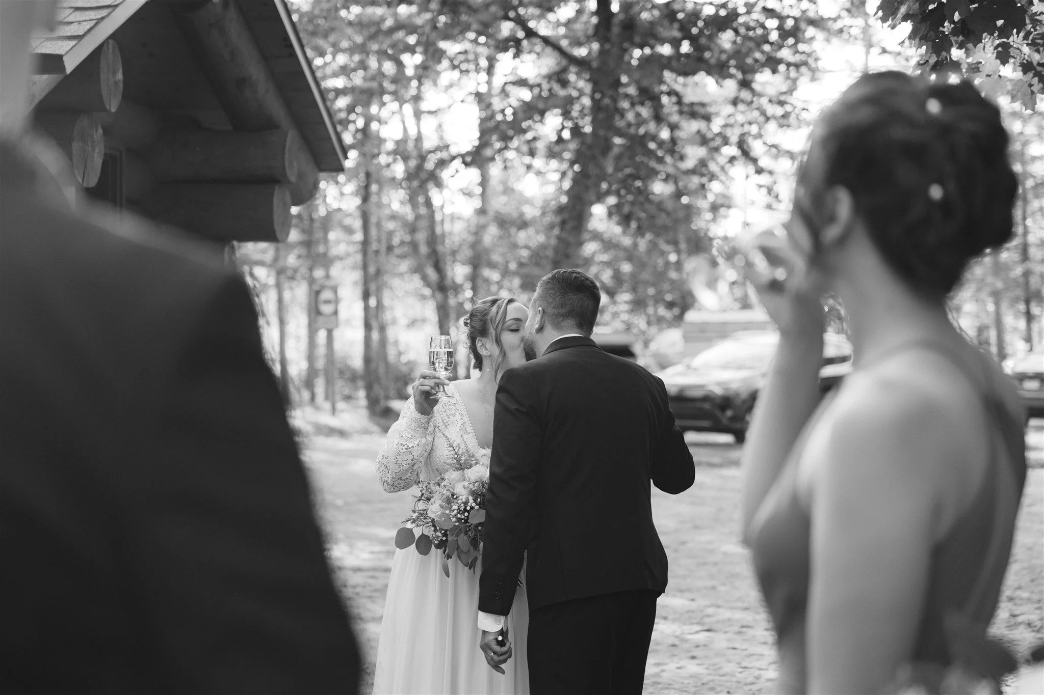 Un couple de mariés s'embrasse durant leur mariage en plein air, entouré d'amis et de famille, avec un décor naturel et des arbres en toile de fond.