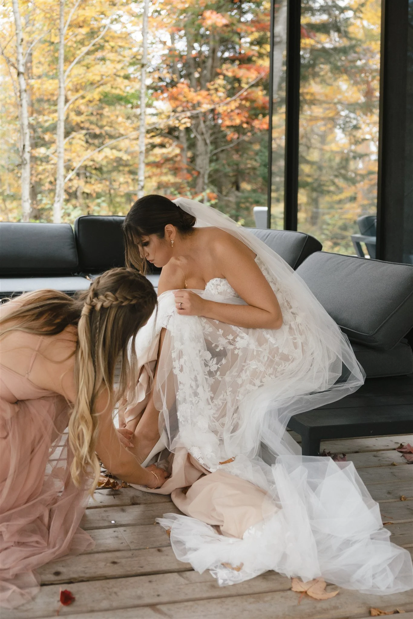 Une mariée en robe de mariage assise sur un canapé, entourée de demoiselles d'honneur en robes roses pâles, dans un environnement intérieur avec de grandes fenêtres donnant sur un paysage d'automne, avec des feuilles mortes au sol.