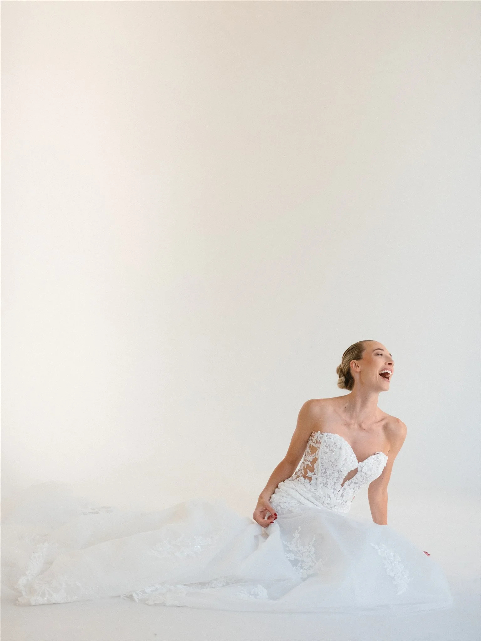 Femme souriante portant une robe de mariée blanche, assise sur le sol blanc dans un intérieur minimaliste.