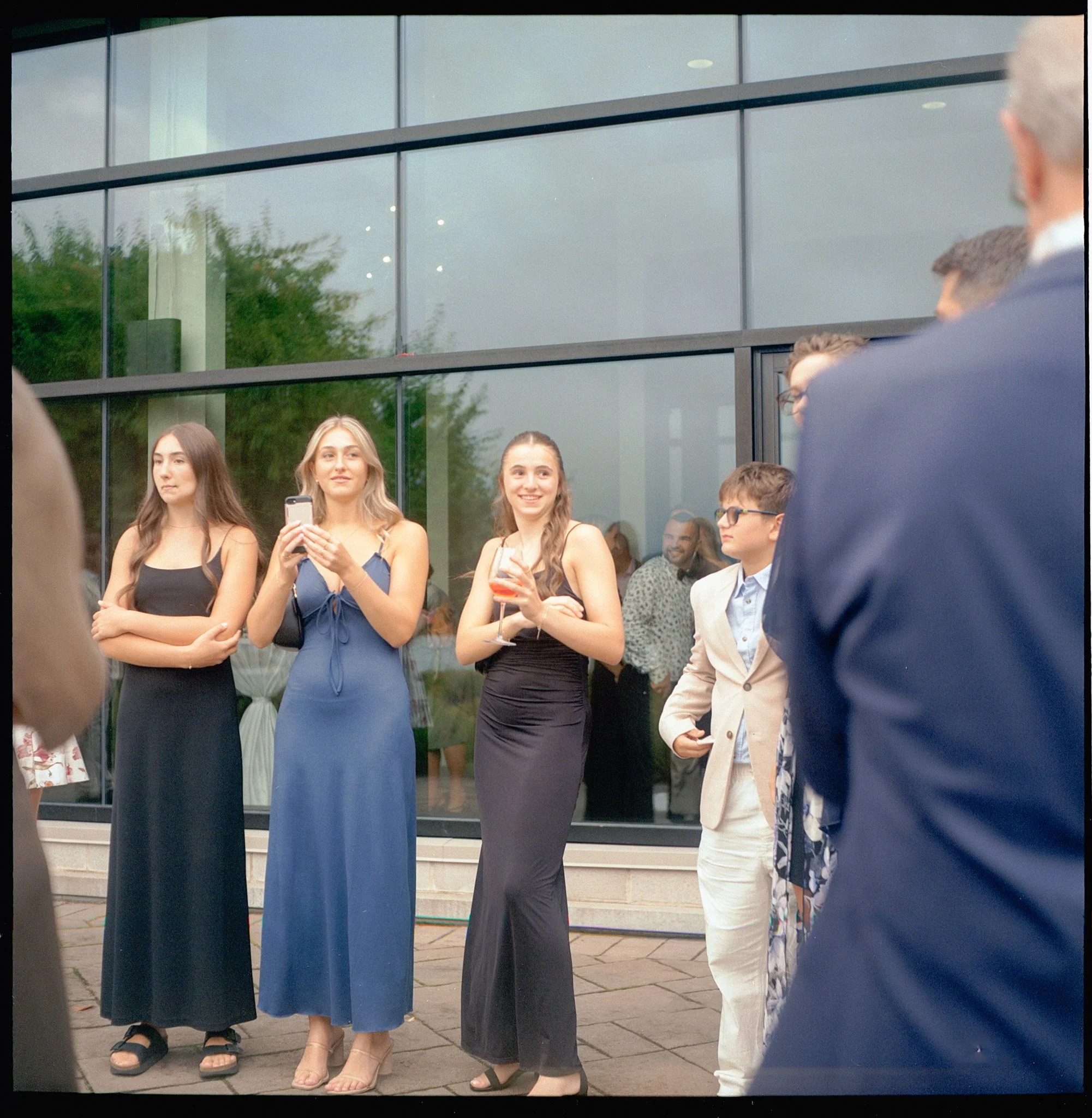 Groupe de jeunes femmes habillées de tenues de soirée noires et bleues, certaines tenant des verres, se tenant devant un bâtiment moderne avec des fenêtres en verre, lors d'un rassemblement extérieur.