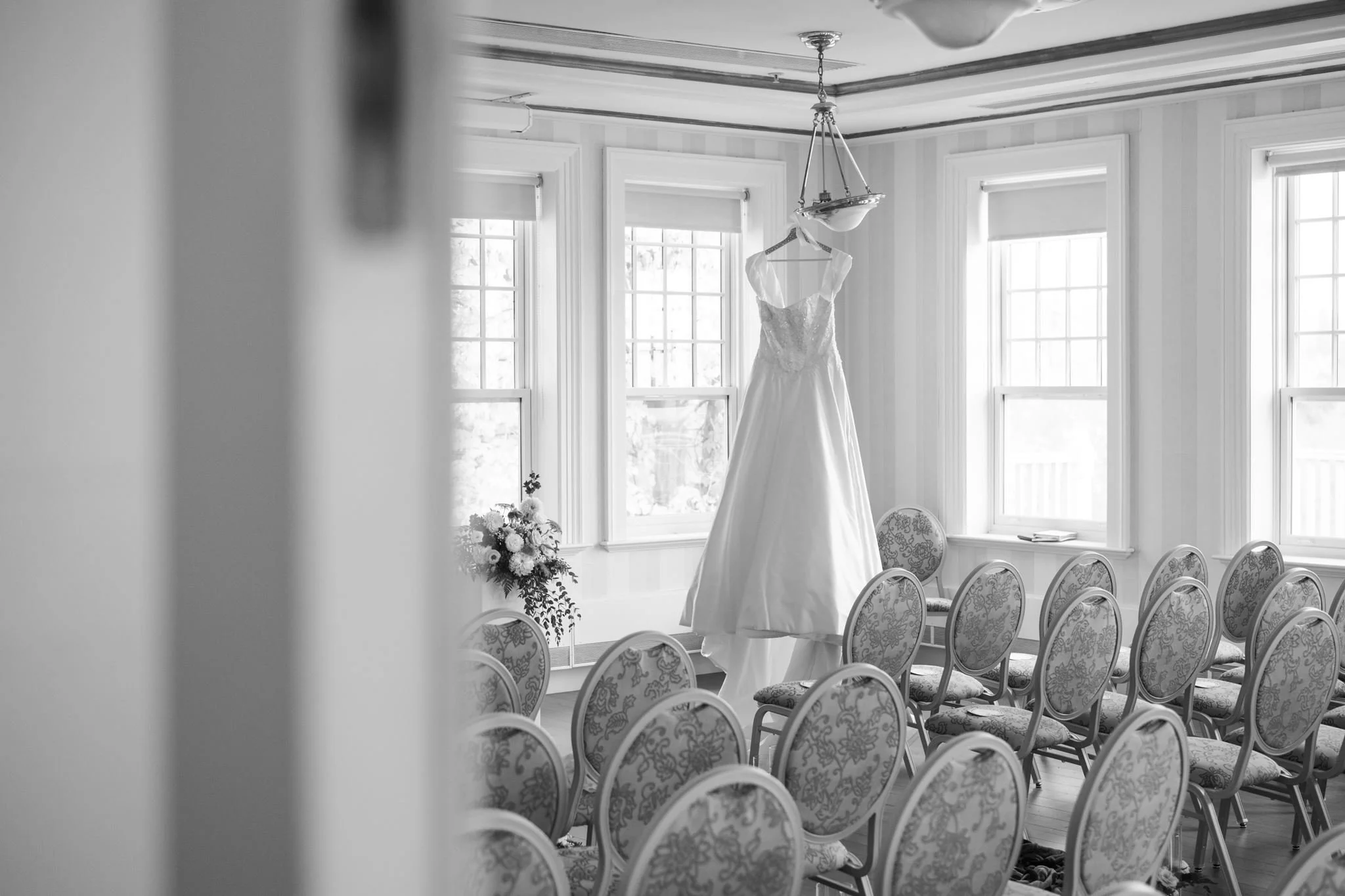 Une robe de mariée suspendue au plafond d'une pièce bien éclairée par de grandes fenêtres, avec des chaises alignées en rangée devant. L'atmosphère semble être une salle de préparation pour un mariage.