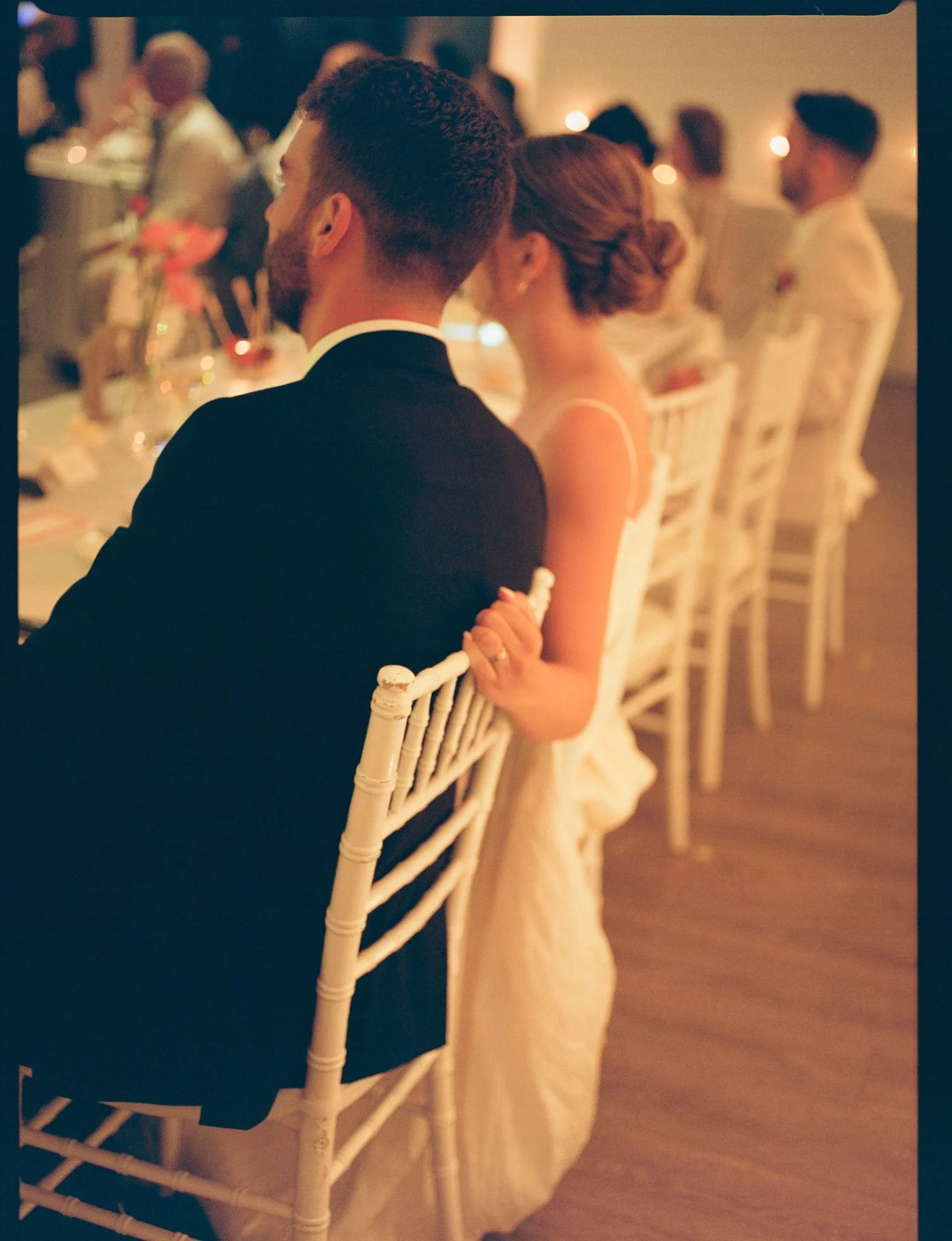 Gros plan sur un couple assis à une table lors d'un dîner de mariage ou d'un banquet. La scène est chaleureuse avec une lumière tamisée et des personnes en arrière-plan.