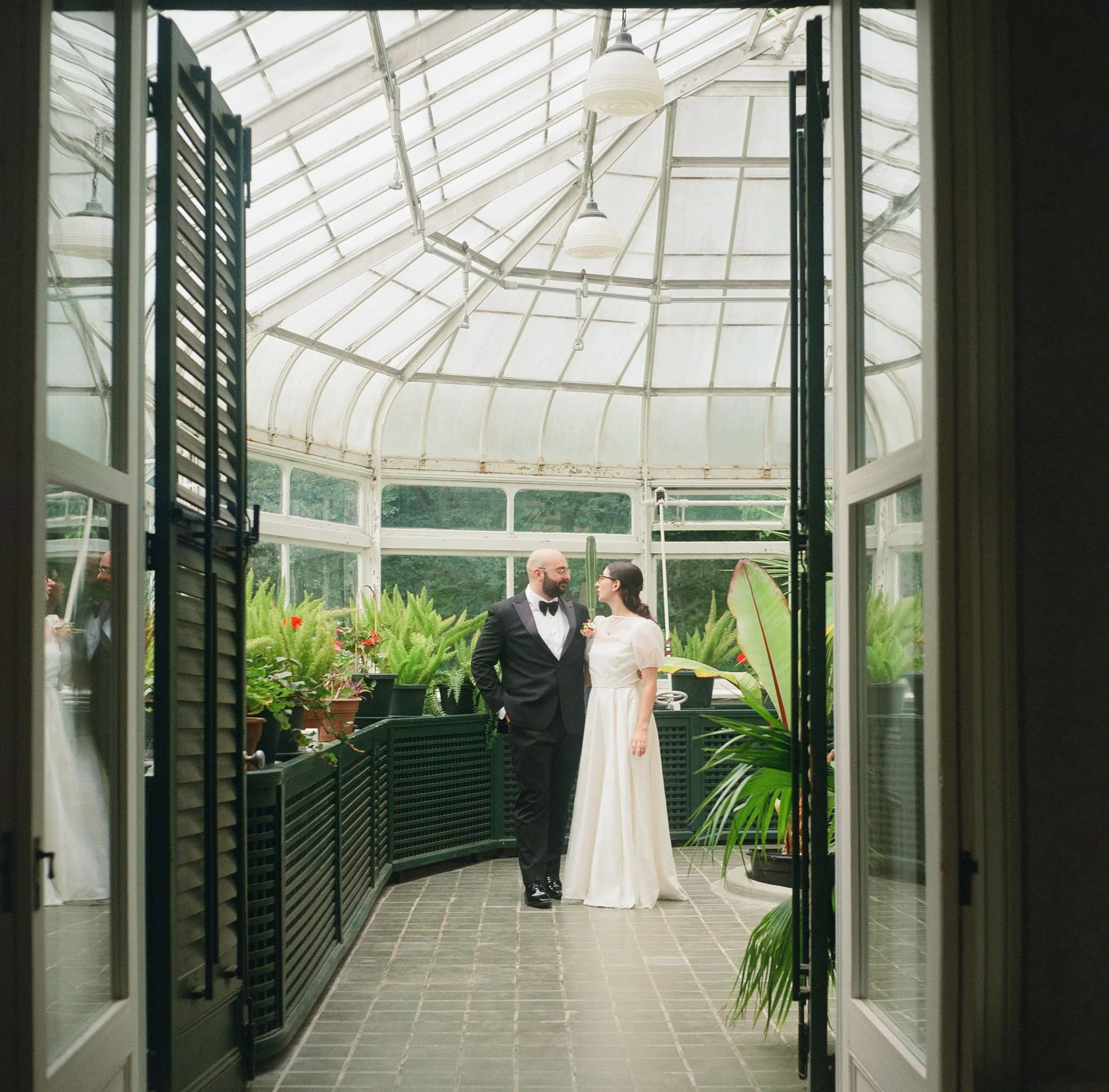 Un couple en costume habillé pour un mariage, se tenant dans une serre ou un jardin d'intérieur, entouré de plantes vertes.
