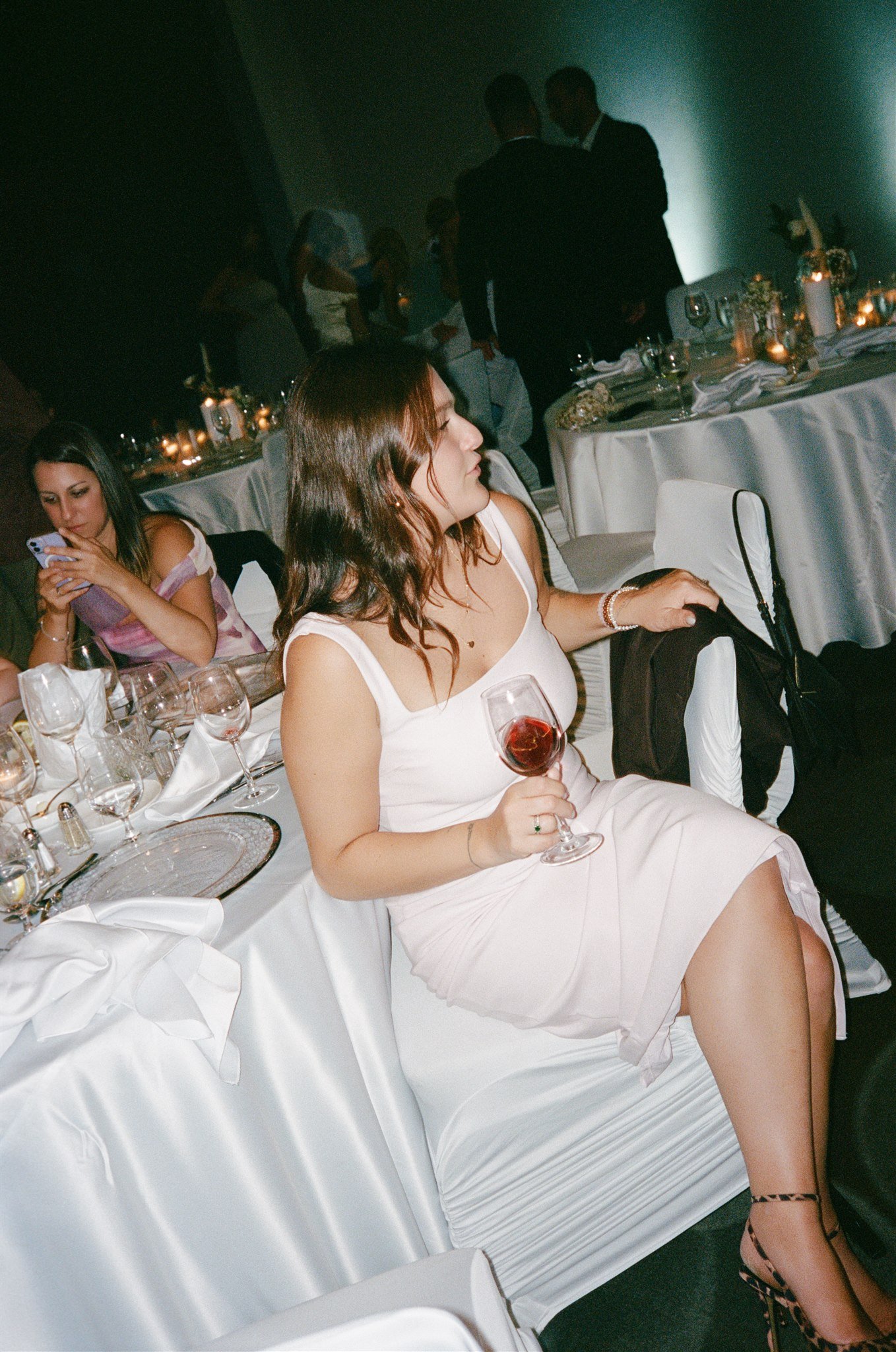 Une femme assise à une table de banquet, tenant un verre de vin rouge, portant une robe blanche, avec des chaussures à talons à motif léopard. Une autre femme à côté regarde son téléphone. Le fond montre des personnes en costume dans une salle décoré