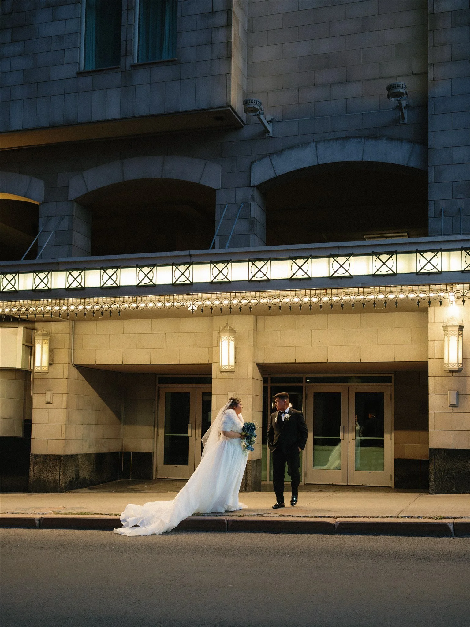 Un couple de mariés en costume et robe de mariée, se tenant la main, devant un bâtiment en pierre avec des lumières décoratives, en soirée.