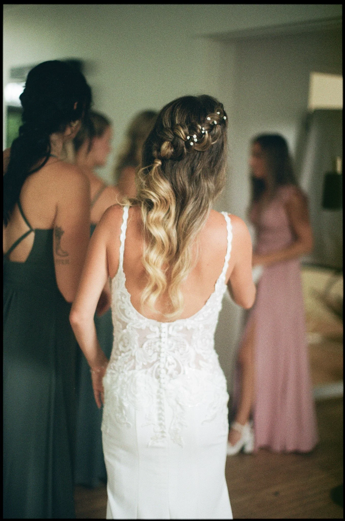 Une femme en robe de mariée blanche avec des boucles d'oreilles, coiffée d'une tresse décorée de perles, préparée pour un mariage, entourée d'autres femmes dans une pièce.