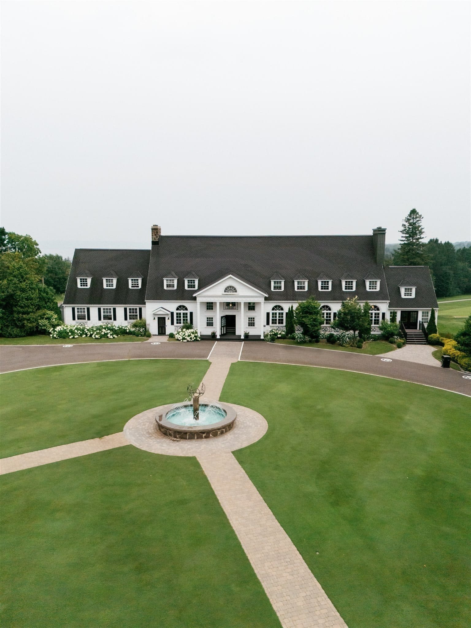 Vue aérienne d’une grande maison blanche au toit noir, entourée d’un jardin soigné, avec une fontaine centrale surmontée d’une sculpture et une allée en pierre menant à l’entrée, captée par un Photographe mariage Québec au Club Royal Québec.