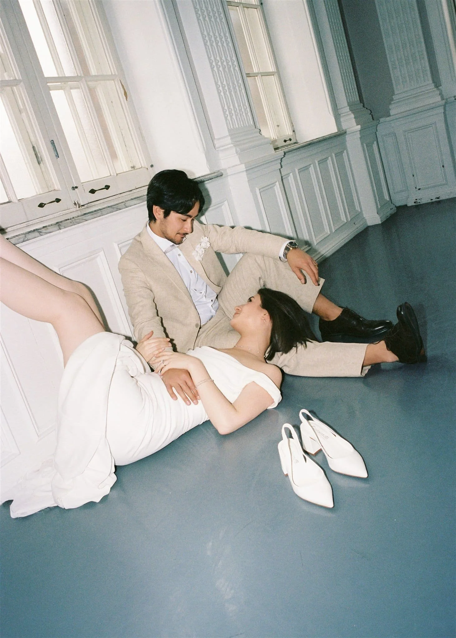 Homme en costume beige et femme en robe blanche avec chaussures à talons, allongés au sol dans une pièce élégante aux murs blancs et grandes fenêtres, se regardant affectueusement, captés par un Photographe mariage Québec.