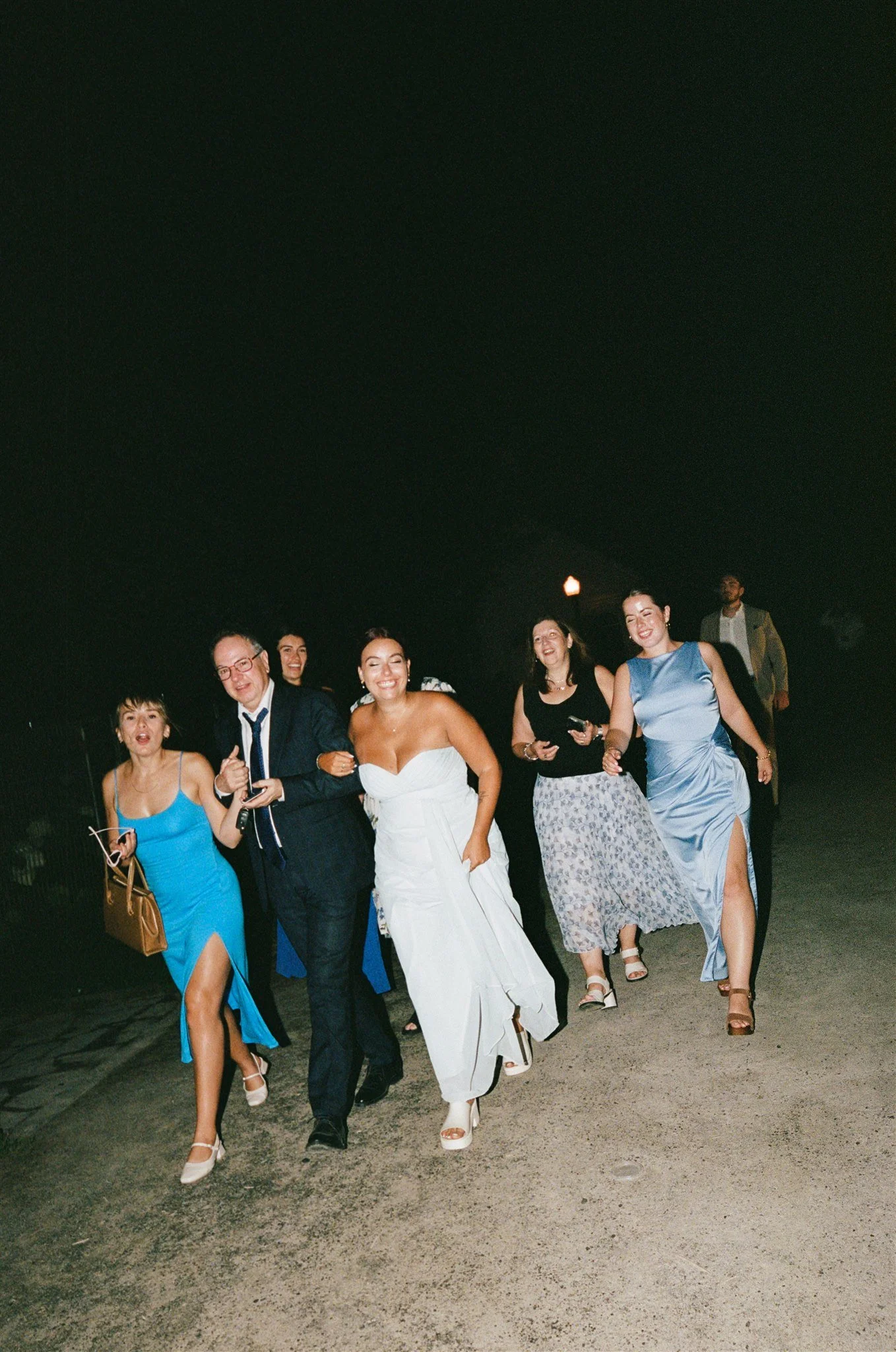 Groupe de personnes habillées élégamment, marchant joyeusement lors d'une célébration la nuit, avec des sourires et une ambiance festive.