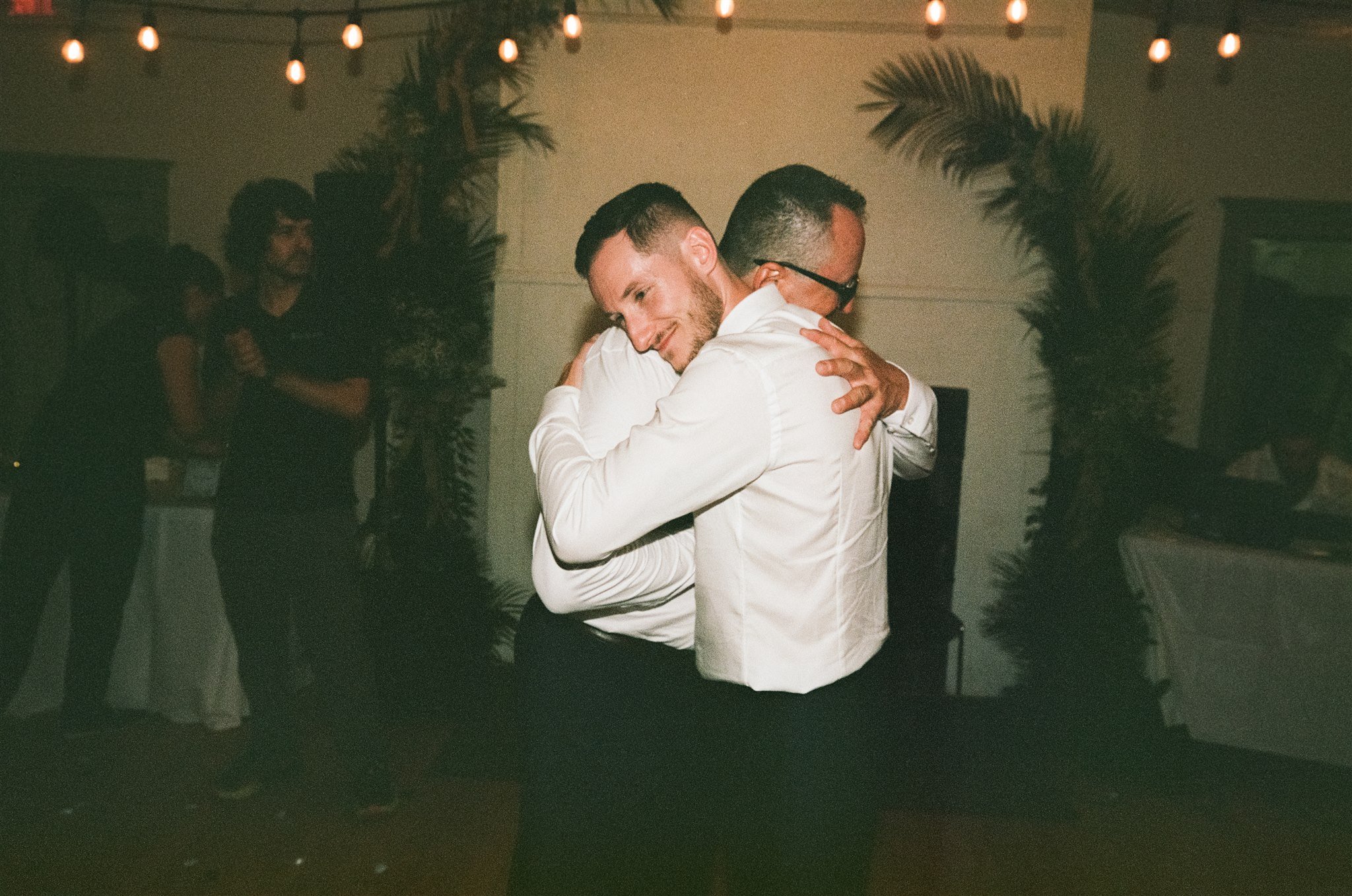 Deux hommes en costume blanc s'embrassent lors d'une danse ou d'une célébration, avec des personnes en arrière-plan et des lumières décoratives.