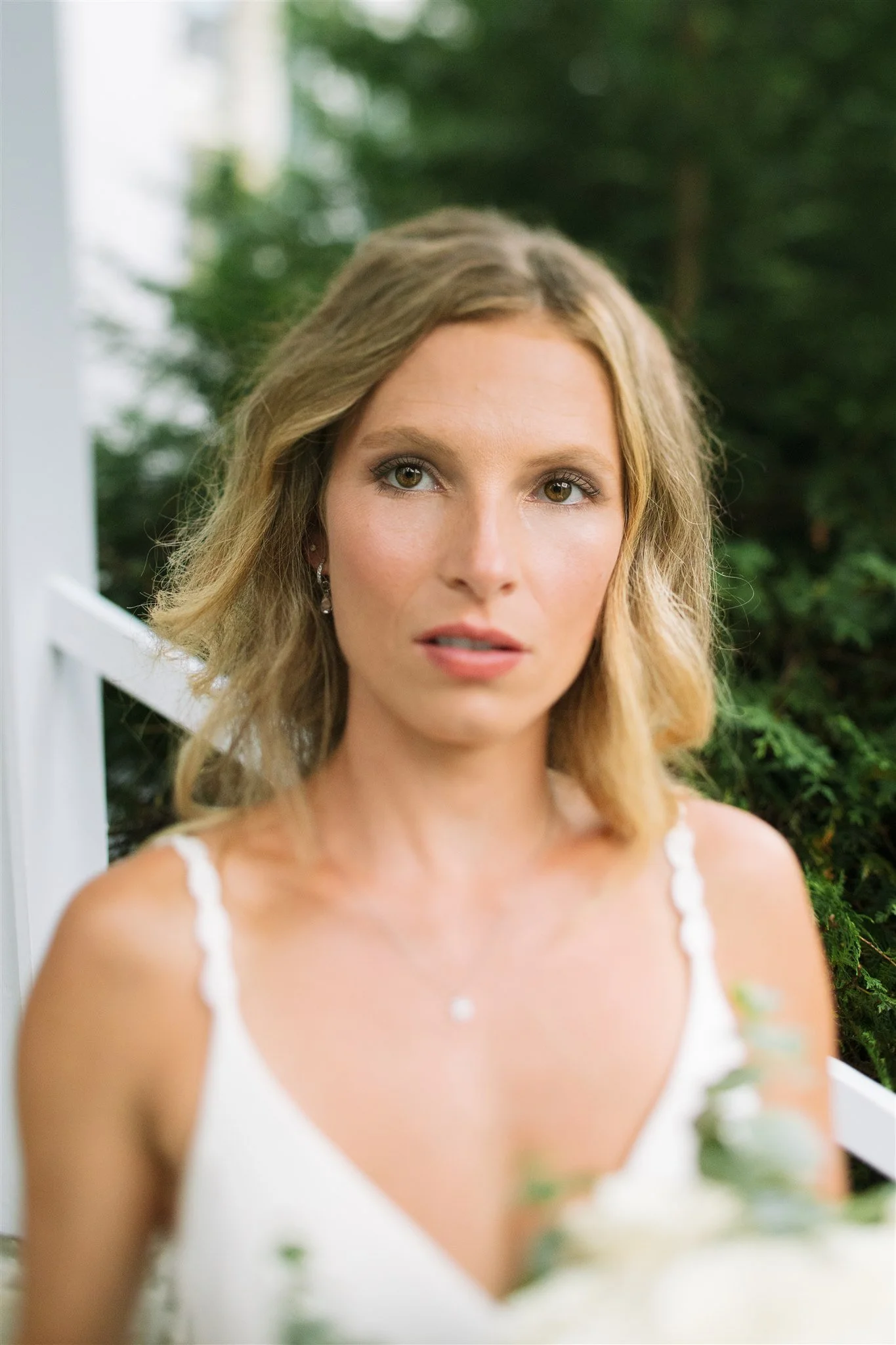 Portrait d'une femme blonde en robe blanche avec des bretelles, debout devant un décor naturel avec des plantes vertes.