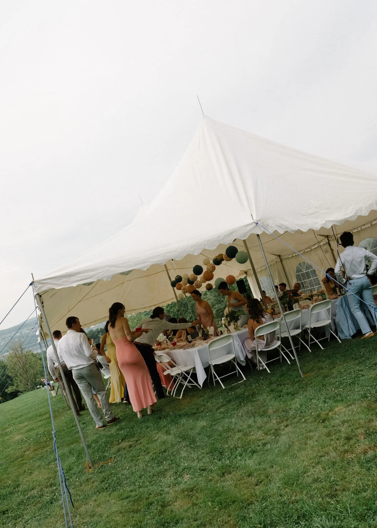 Une fête en plein air sous une grande tente blanche, avec des gens assis et debout autour d'une table, décorée avec des lanternes colorées suspendues et des invités élégants, probablement lors d'un mariage ou d'une réception.