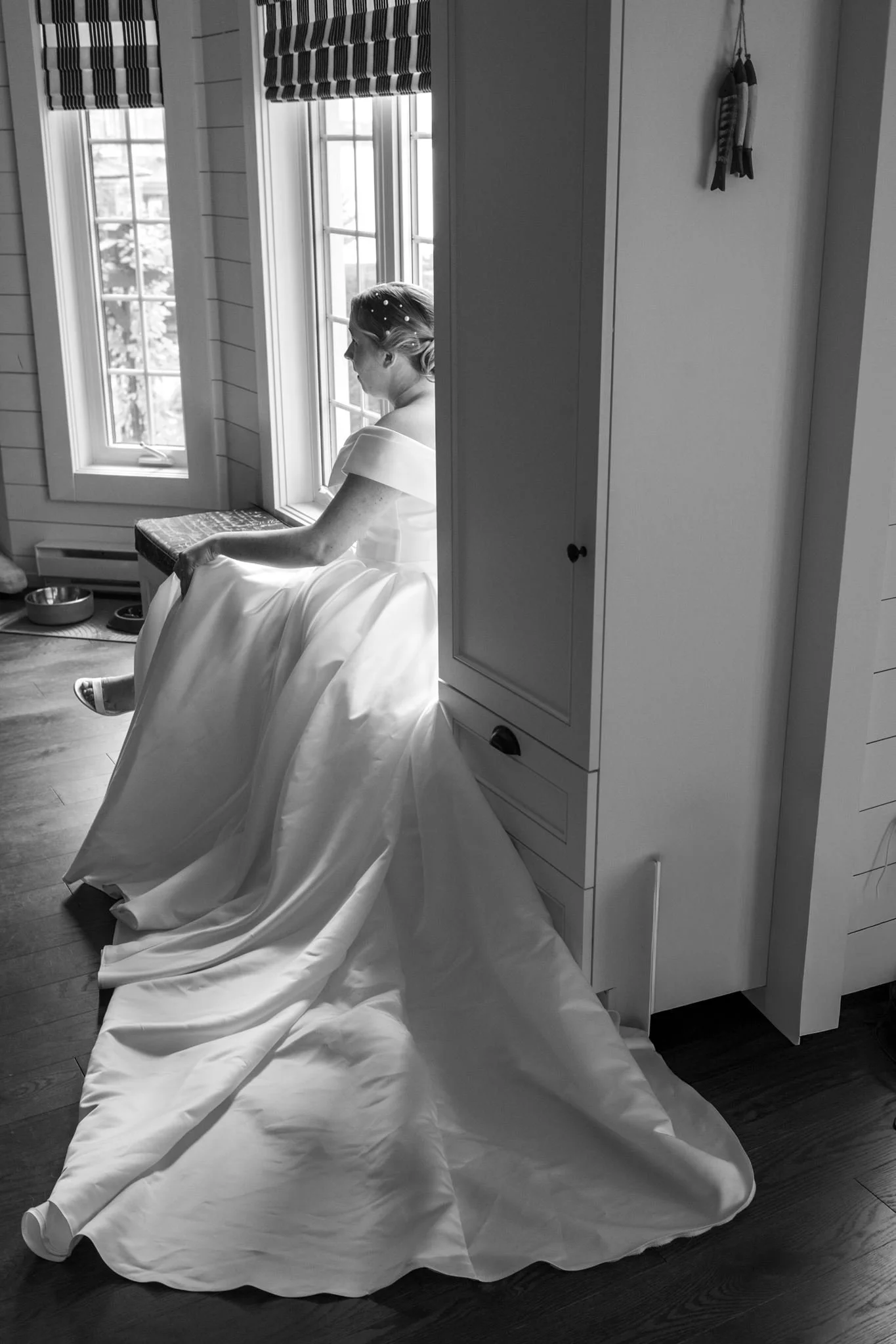 Femme en robe de mariée assise près de fenêtres, dans une pièce en bois, avec un décor simple.