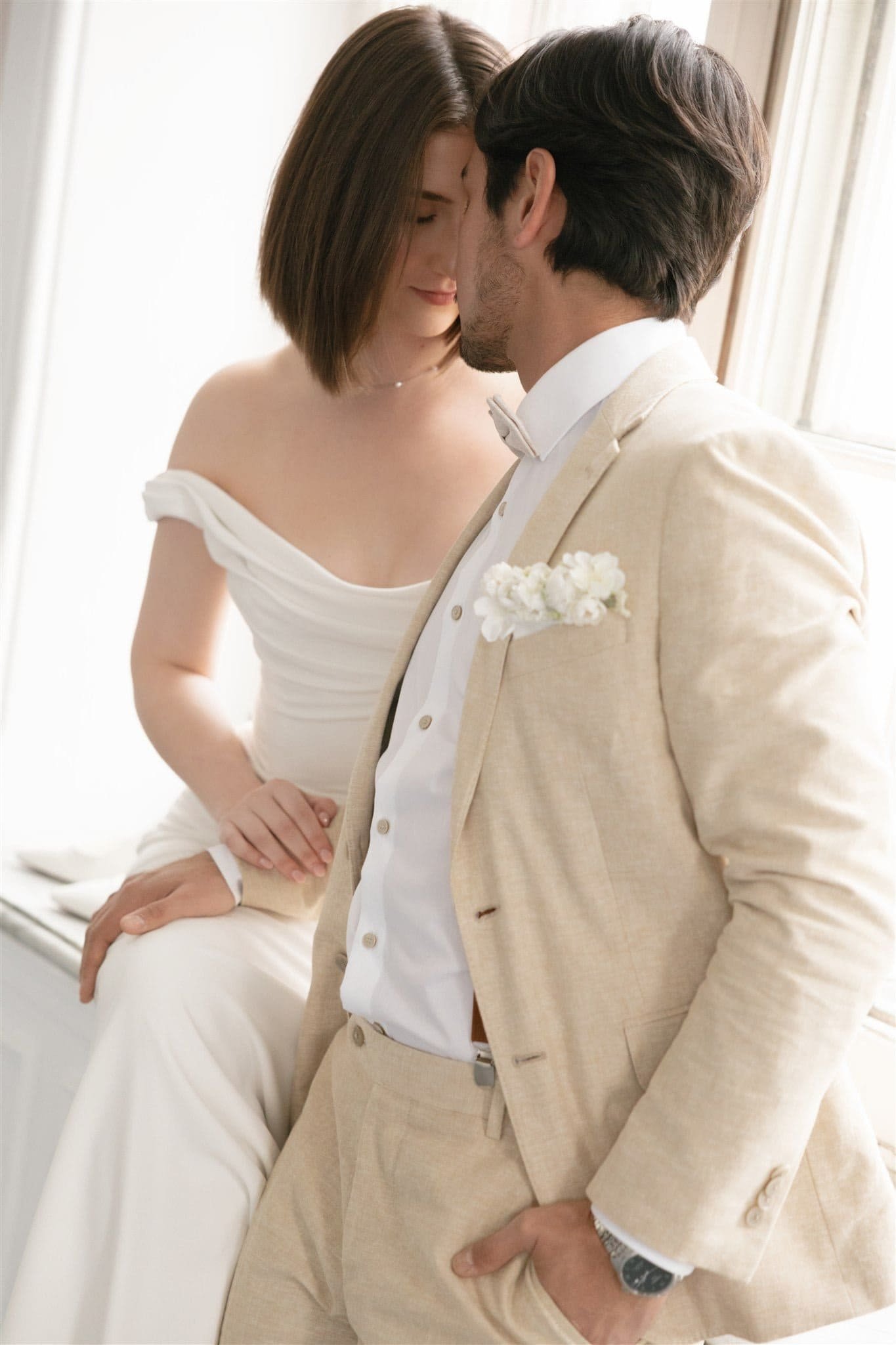Couple en tenue de mariage, la femme assise et l’homme debout, posant tendrement front et nez touchant dans une pièce lumineuse, captés par un Photographe mariage Québec.
