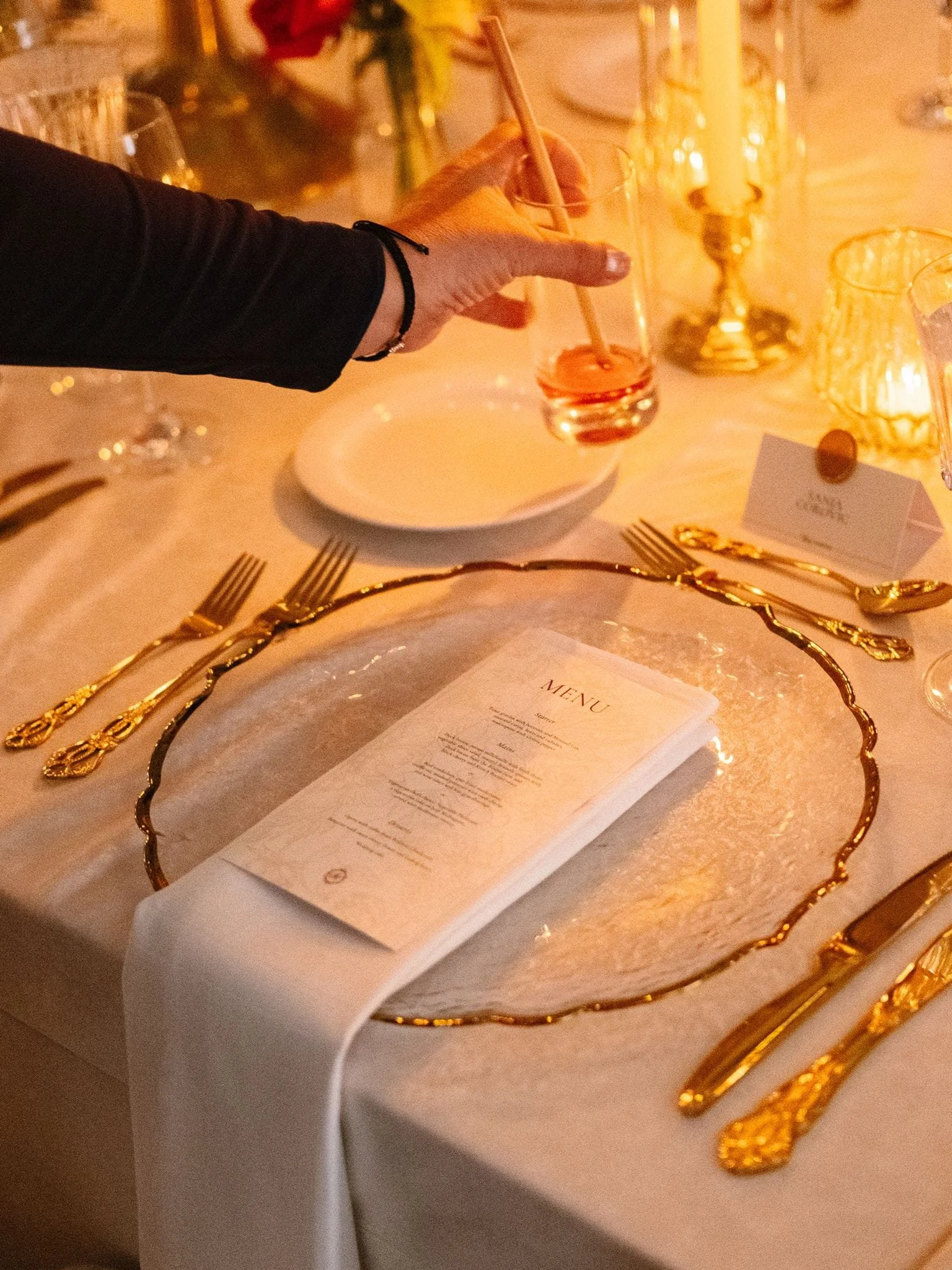 Une personne sert un verre avec une paille dans un cadre de dîner, avec une assiette, des fourchettes dorées, un menu, des bougies et une décoration florale élégante.