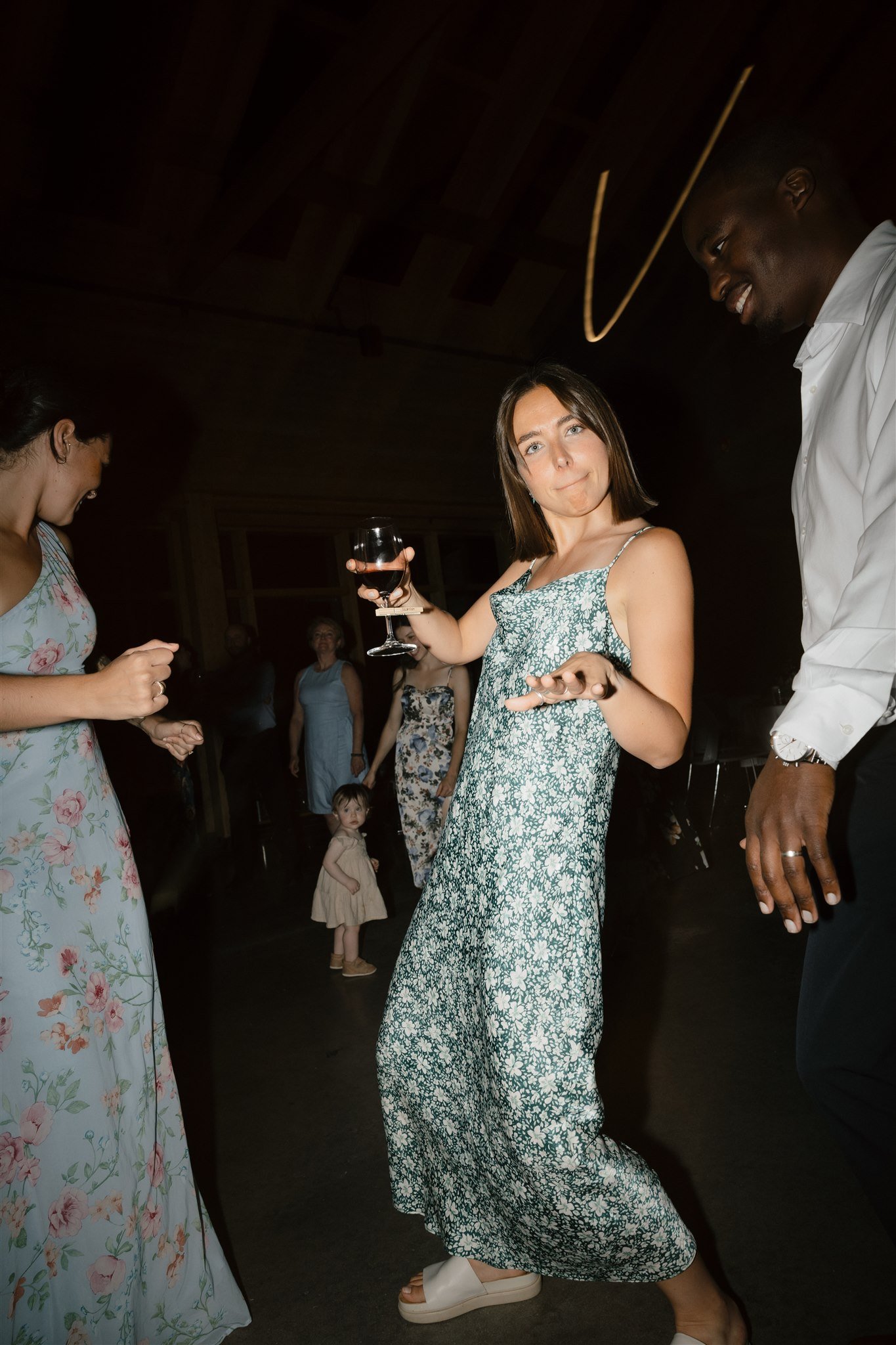 Une femme en robe à fleurs tenant un verre de vin rouge, entourée de plusieurs personnes lors d'une fête ou d'un événement intérieur.