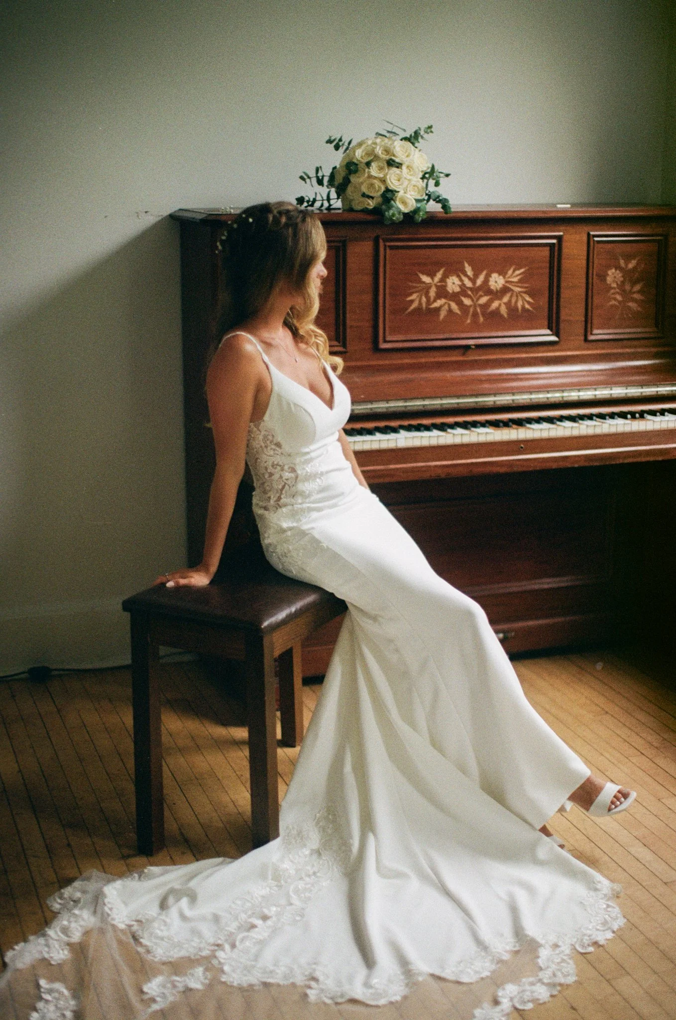 Une femme en robe de mariage blanche assise sur un banc à côté d'un piano en bois, avec un bouquet de roses blanches sur le piano.