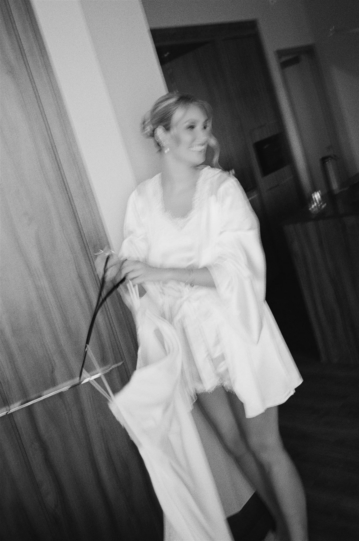 Une femme souriante portant une robe blanche en satin, debout près d'une armoire en bois dans une pièce intérieure.