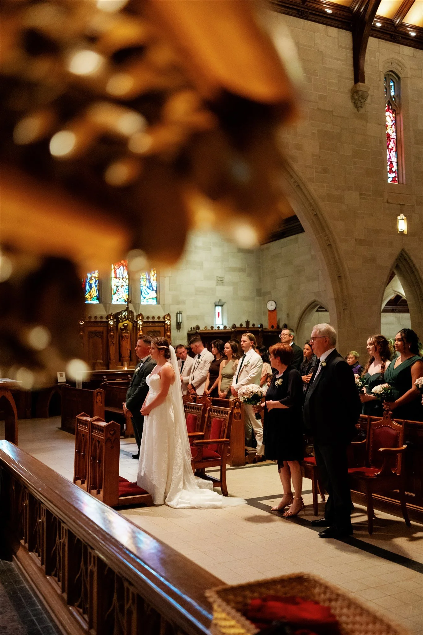 Une cérémonie de mariage dans une église avec des personnes debout, dont la mariée en robe blanche. La scène se déroule à l'intérieur d'une église avec des vitraux colorés et des bancs en bois.
