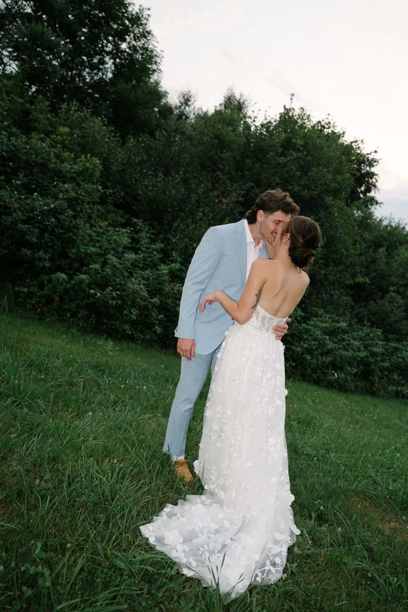 Un couple en robe de mariée et costume se tient dans un jardin verdoyant, s'apprêtant à s'embrasser. La scène est en plein air, avec des arbres en arrière-plan et un ciel nuageux.