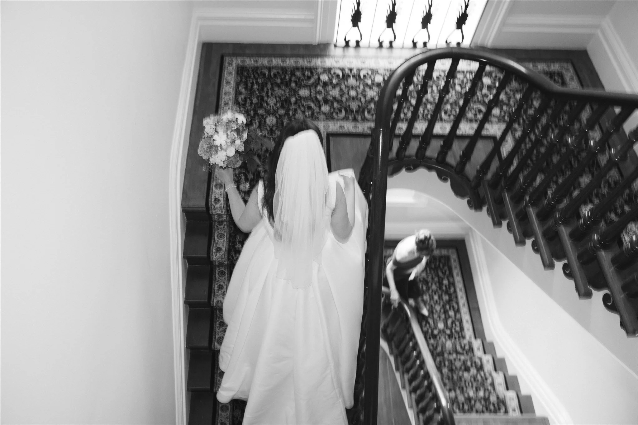 Une femme portant une robe blanche et un voile de mariée descend un escalier en bois dans une maison élégante. Un bouquet de fleurs est placé sur une console à côté de l'escalier.