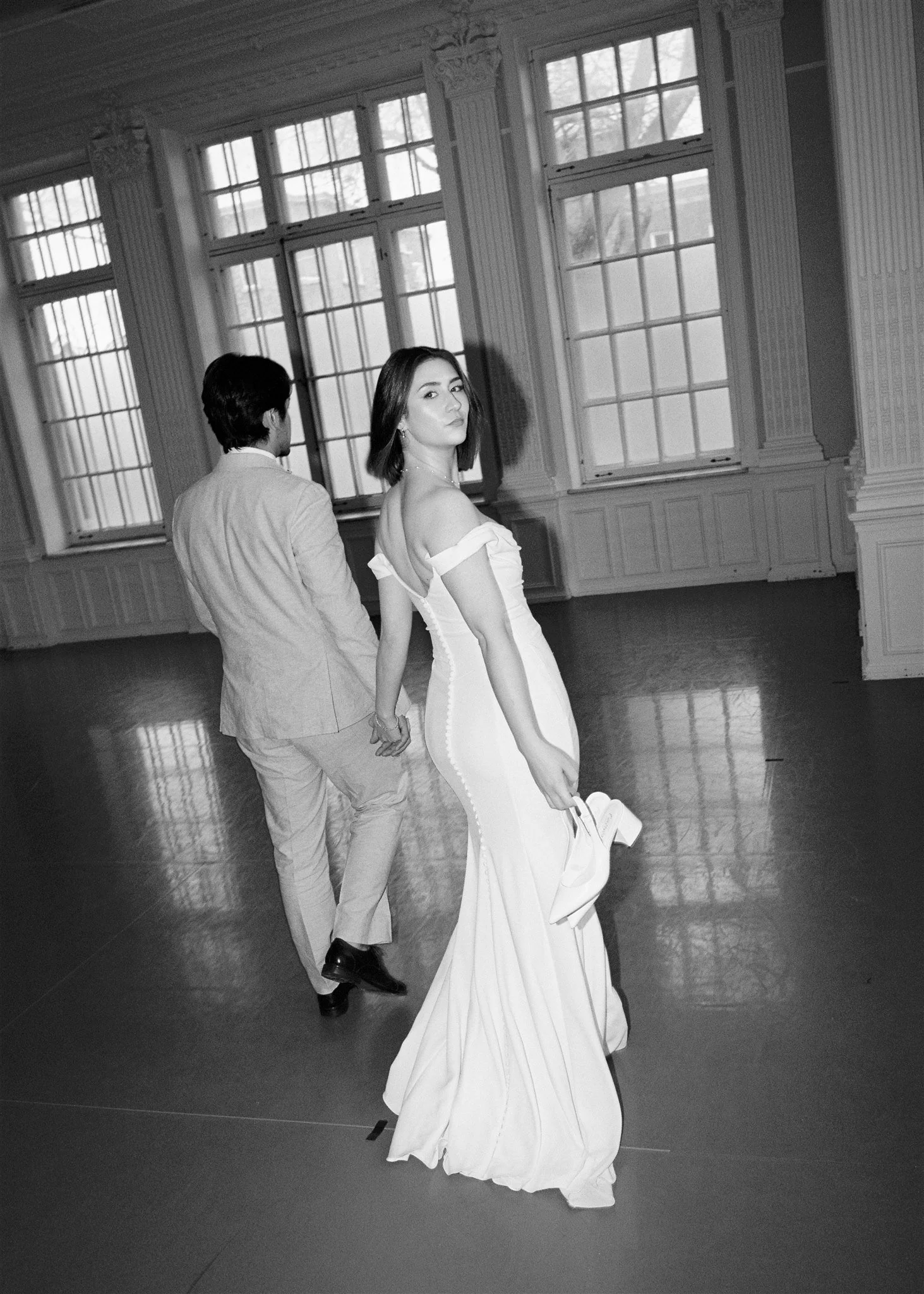 Une femme en robe de bal blanche et un homme en costume noir dans une pièce élégante avec de grandes fenêtres.