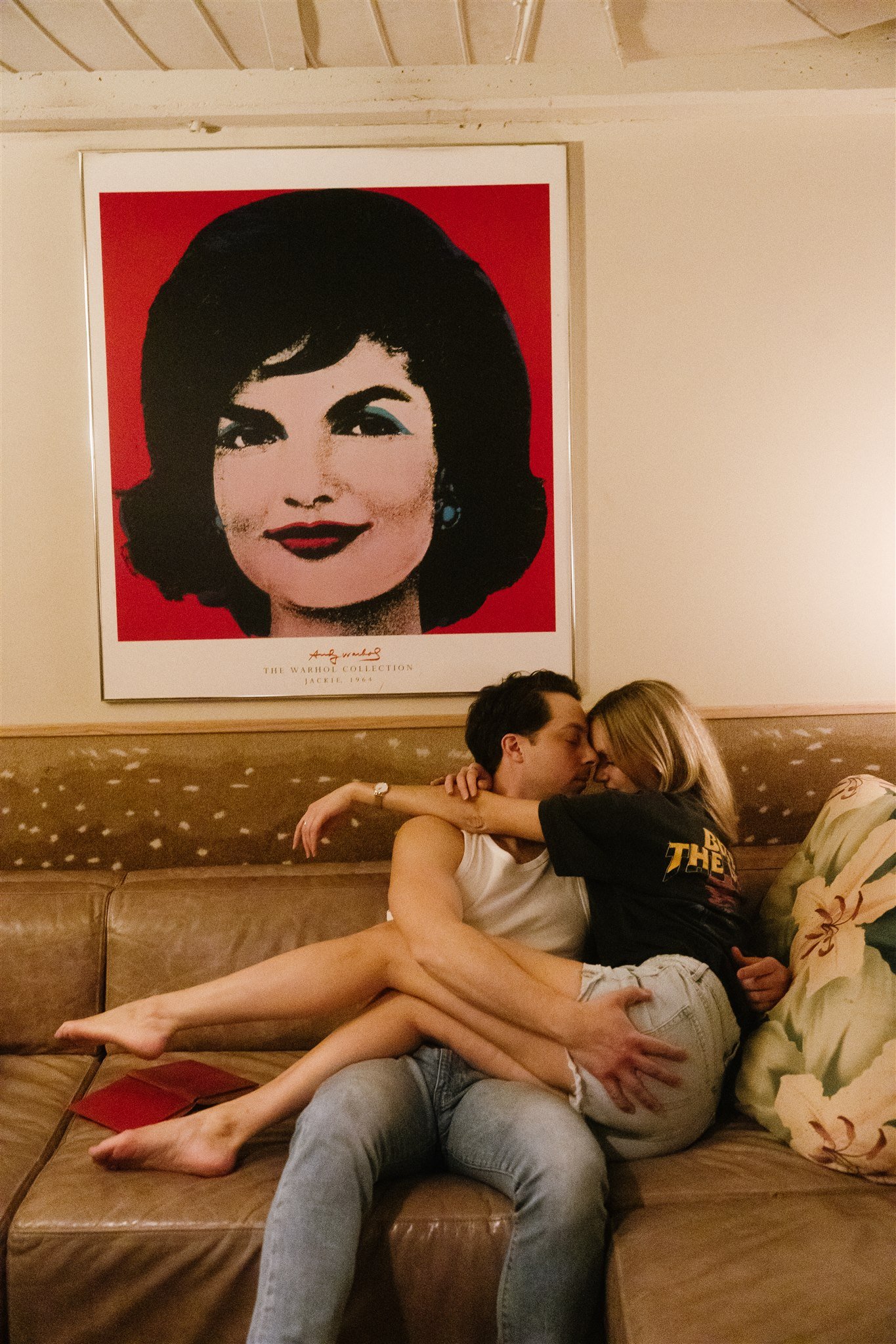 Une photo de deux personnes assises sur un canapé, s'embrassant tendrement, dans une pièce chaleureuse avec un tableau de Marilyn Monroe en fond.