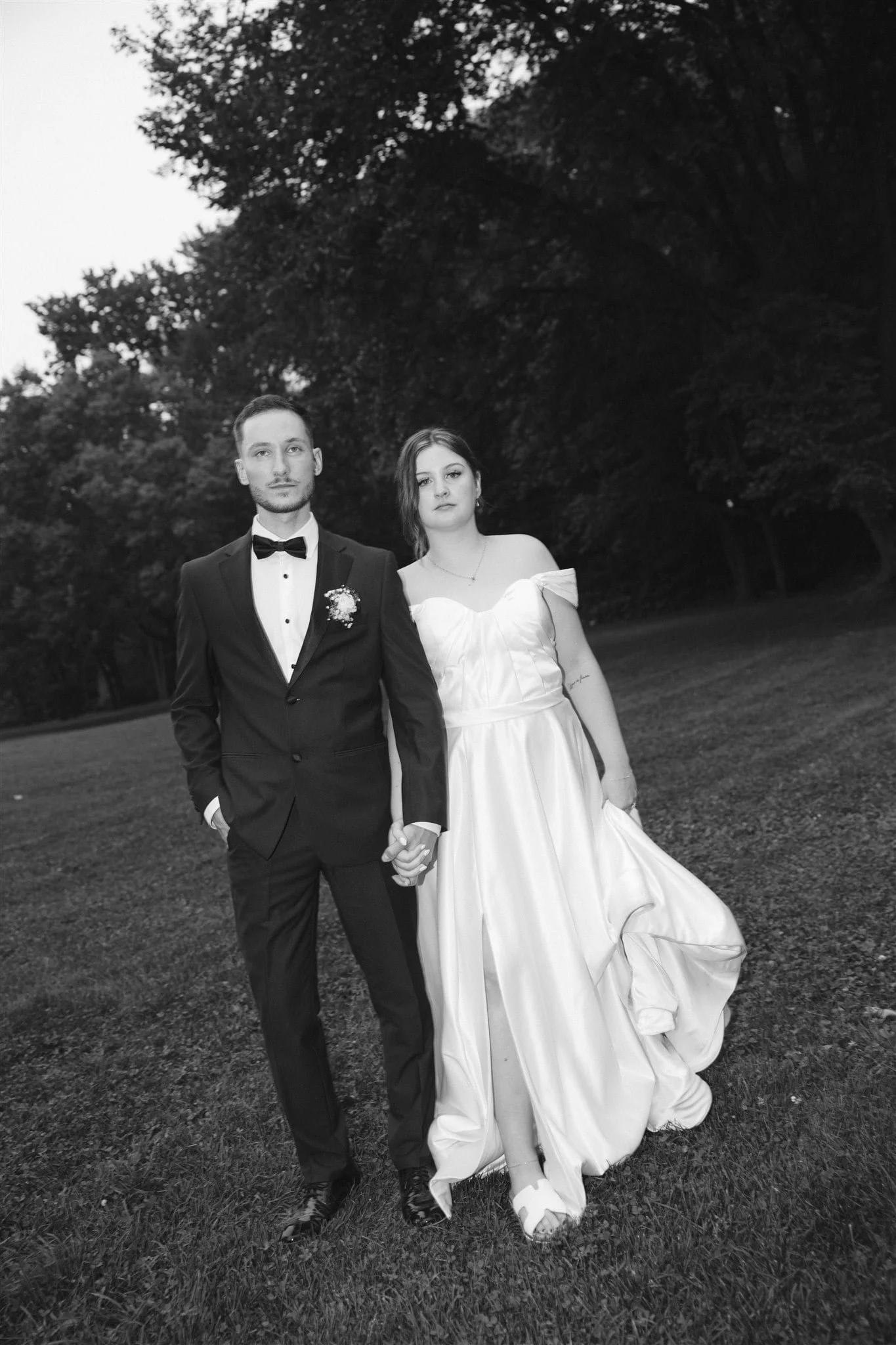 Couple en tenue de mariage marchant dans un parc, la mariée en robe blanche élégante et le marié en costume noir avec nœud papillon, en noir et blanc, captés par un Photographe mariage Québec à l’Hôtel des Premières Nations.