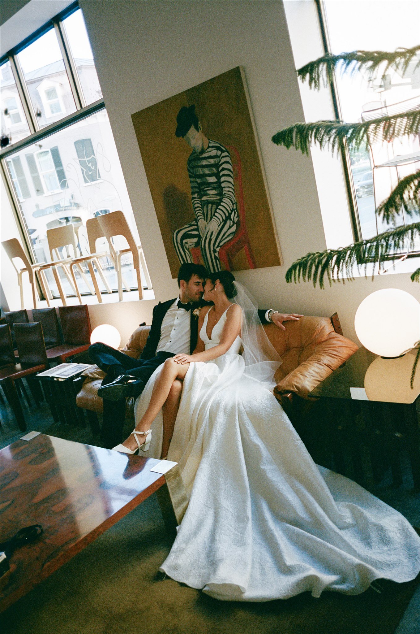 Un couple en mariage assis sur un canapé dans un intérieur moderne avec une peinture abstraite sur le mur derrière eux. La mariée porte une robe blanche et des talons blancs, le marié porte un costume noir avec une chemise blanche et un nœud papillon