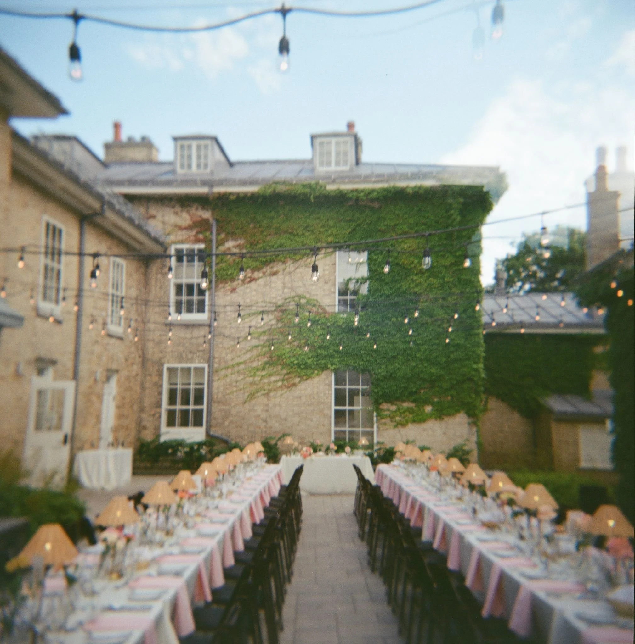 Une longue table dressée avec des nappes roses, des assiettes, des verres et des chaises, dans un espace extérieur pour un événement ou une fête, avec des guirlandes lumineuses suspendues au-dessus.