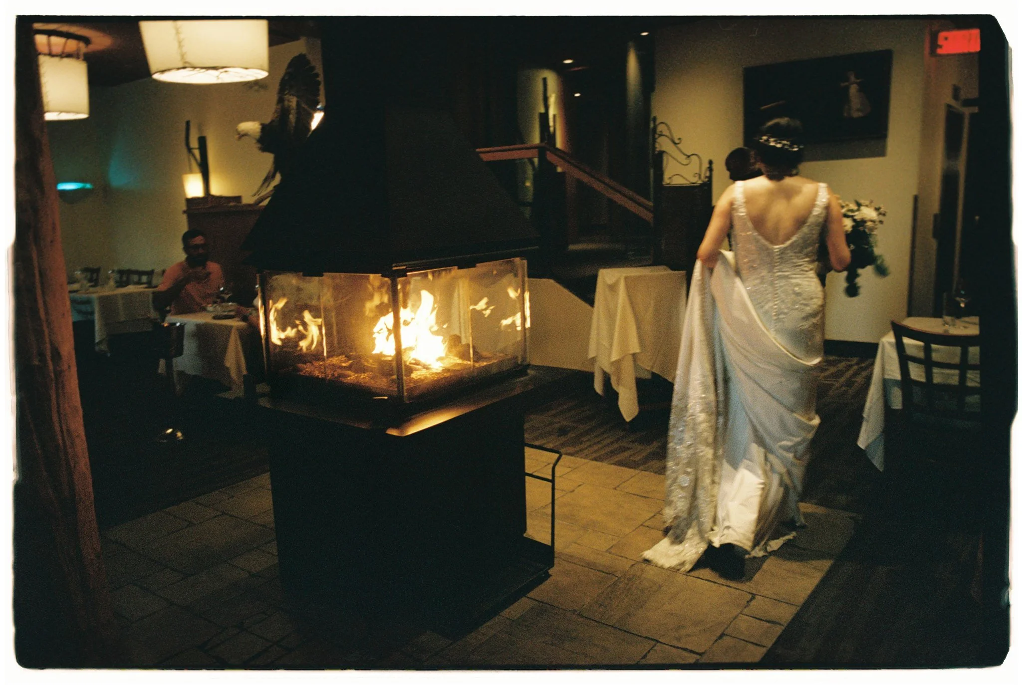 Une scène d'intérieur dans un restaurant avec une cheminée allumée au centre et deux femmes en robe de mariée marchant côte à côte, une à gauche assise à une table et une à droite portant un bouquet.