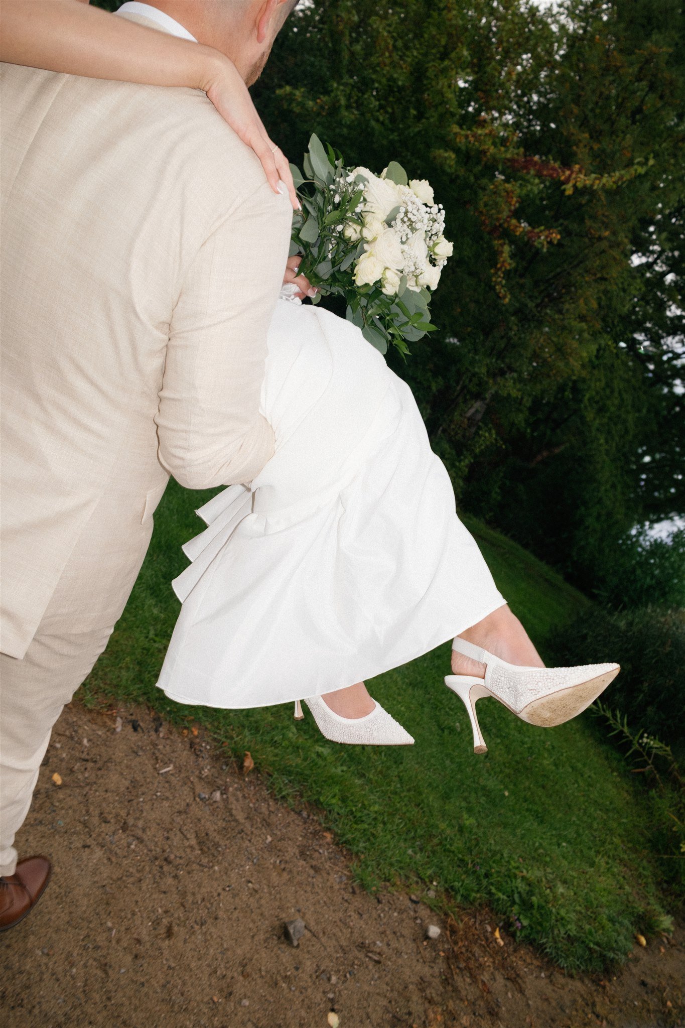 Un couple lors d'un mariage, la femme porte une robe blanche et des talons hauts blancs, la femme porte des chaussures blanches à semelle en bois. La femme tient un bouquet de fleurs blanches et vertes. Ils sont dans un extérieur avec des arbres en a