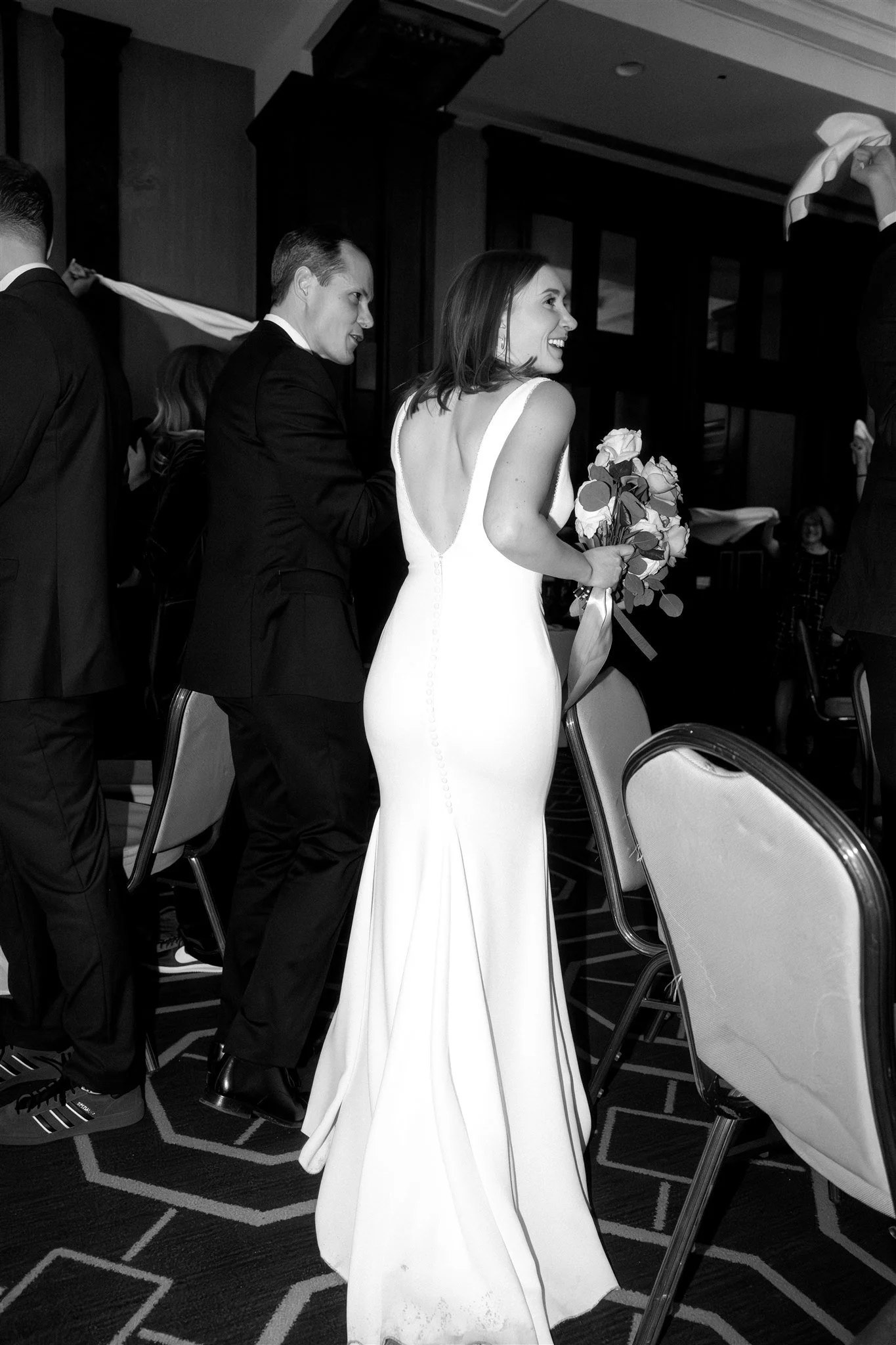 Une femme en robe de mariage souriante, tenant un bouquet de fleurs, assise lors d'une cérémonie ou d'une réception, entourée d'hommes en costume.