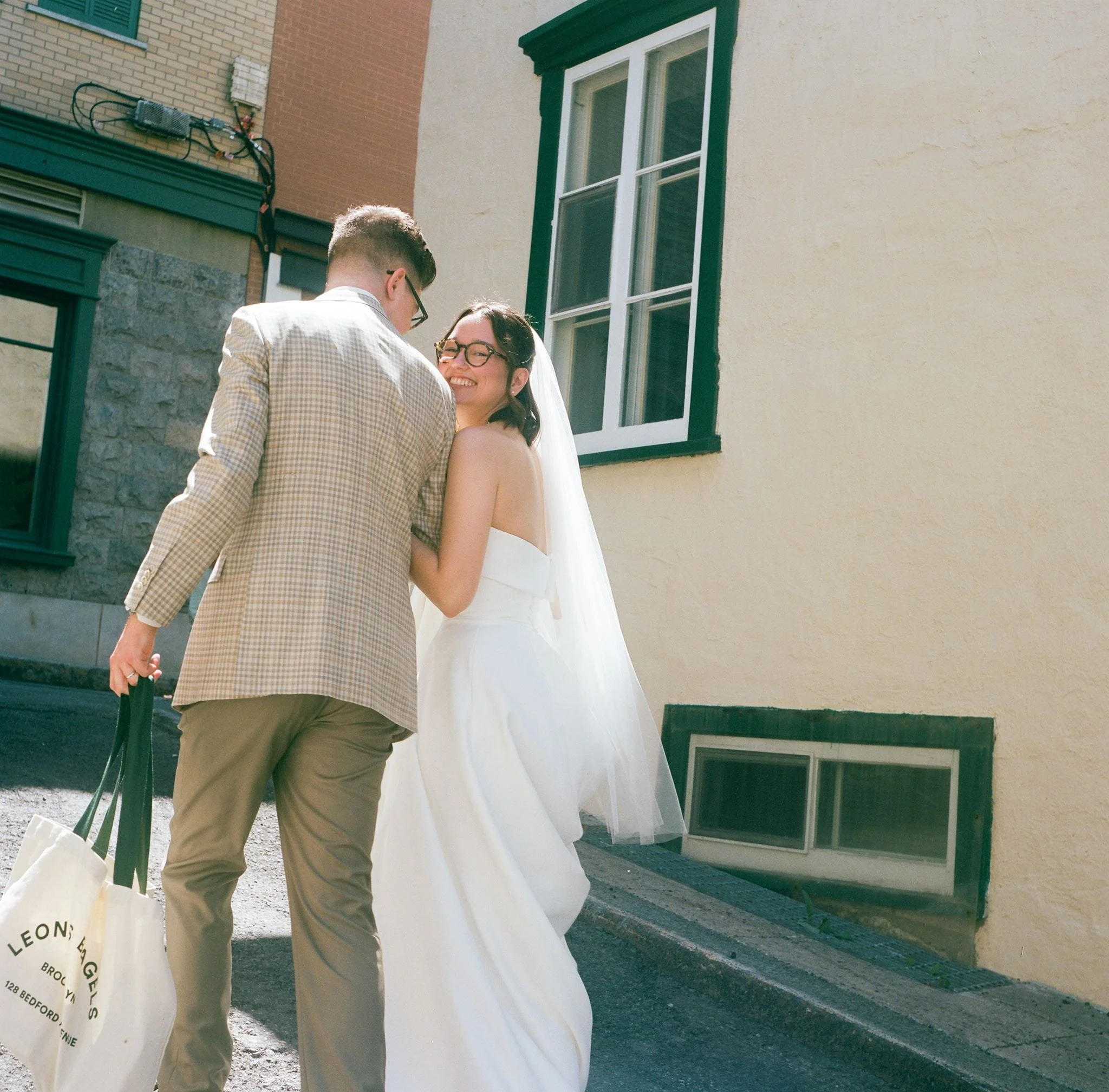 Un homme et une femme se tiennent dans une interaction joyeuse devant un bâtiment avec une fenêtre à cadre vert. Il porte une veste en tartan et des lunettes, elle porte une robe de mariage et des lunettes, souriant. La scène semble être une célébrat
