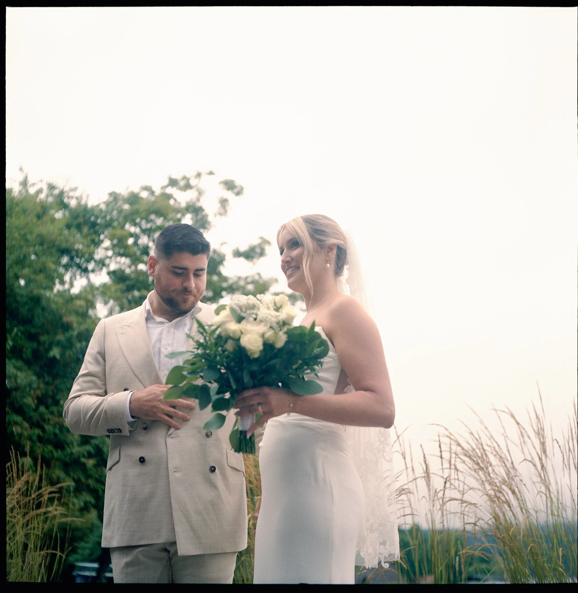Un couple de mariage en costume clair, la femme en robe blanche, échangeant un bouquet de fleurs dans un cadre naturel avec des champs et des arbres en arrière-plan.
