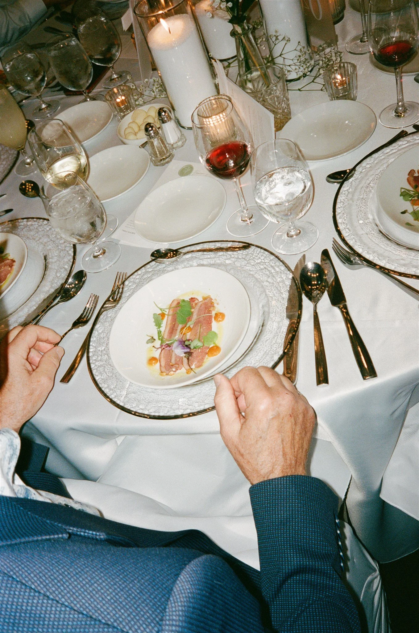 Une table de dîner élégante avec des assiettes en porcelaine, des verres à vin rouge, à eau, de l'eau avec glaçons, et des bougies allumées. Deux mains, probablement d'une personne âgée, tiennent une assiette avec une présentation gastronomique. La t