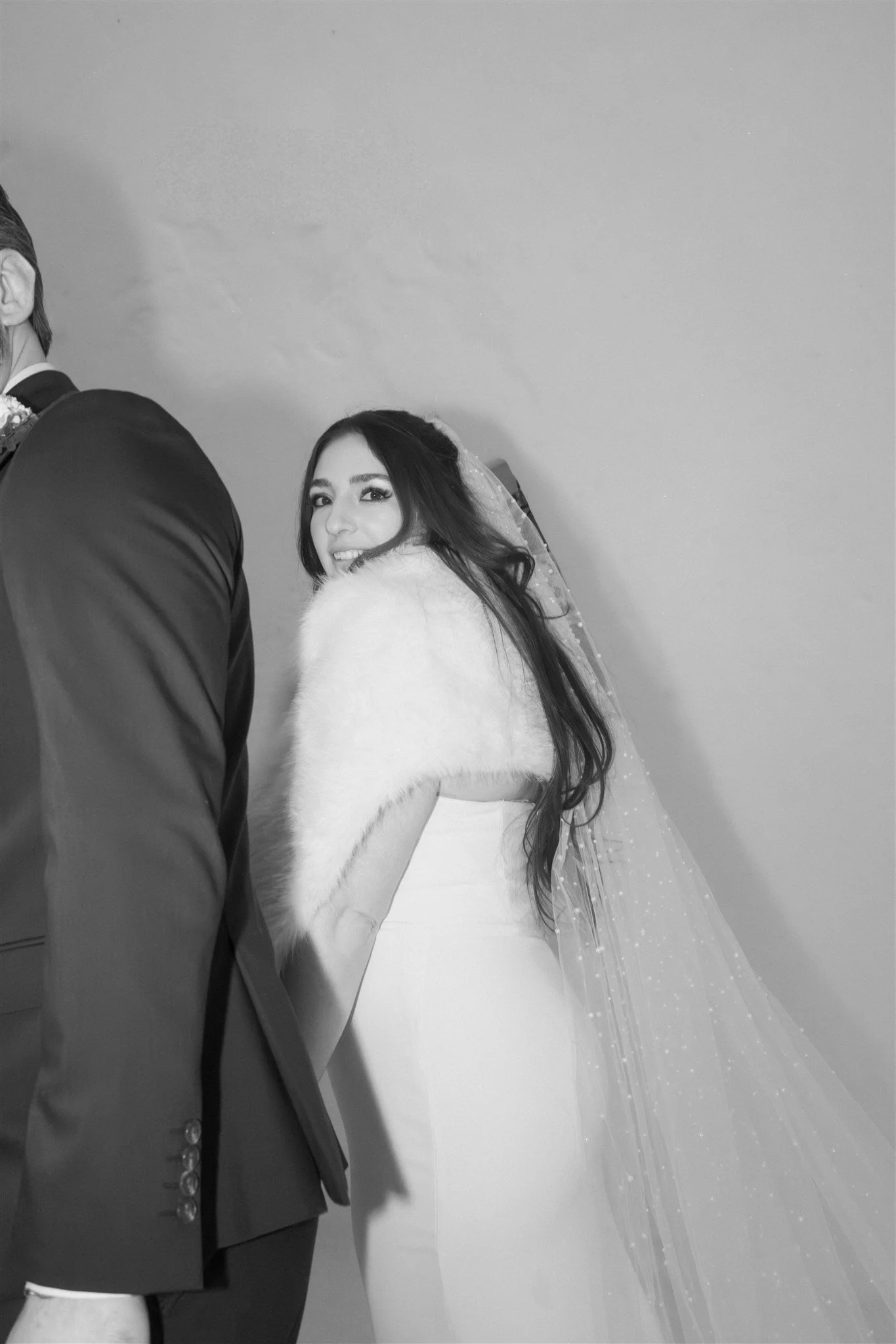 Une femme souriante portant une robe blanche et une étole en fausse fourrure, avec un voile de mariage, posant en compagnie d’un homme en costume à côté d’elle, lors d’un mariage ou d’une cérémonie spéciale.